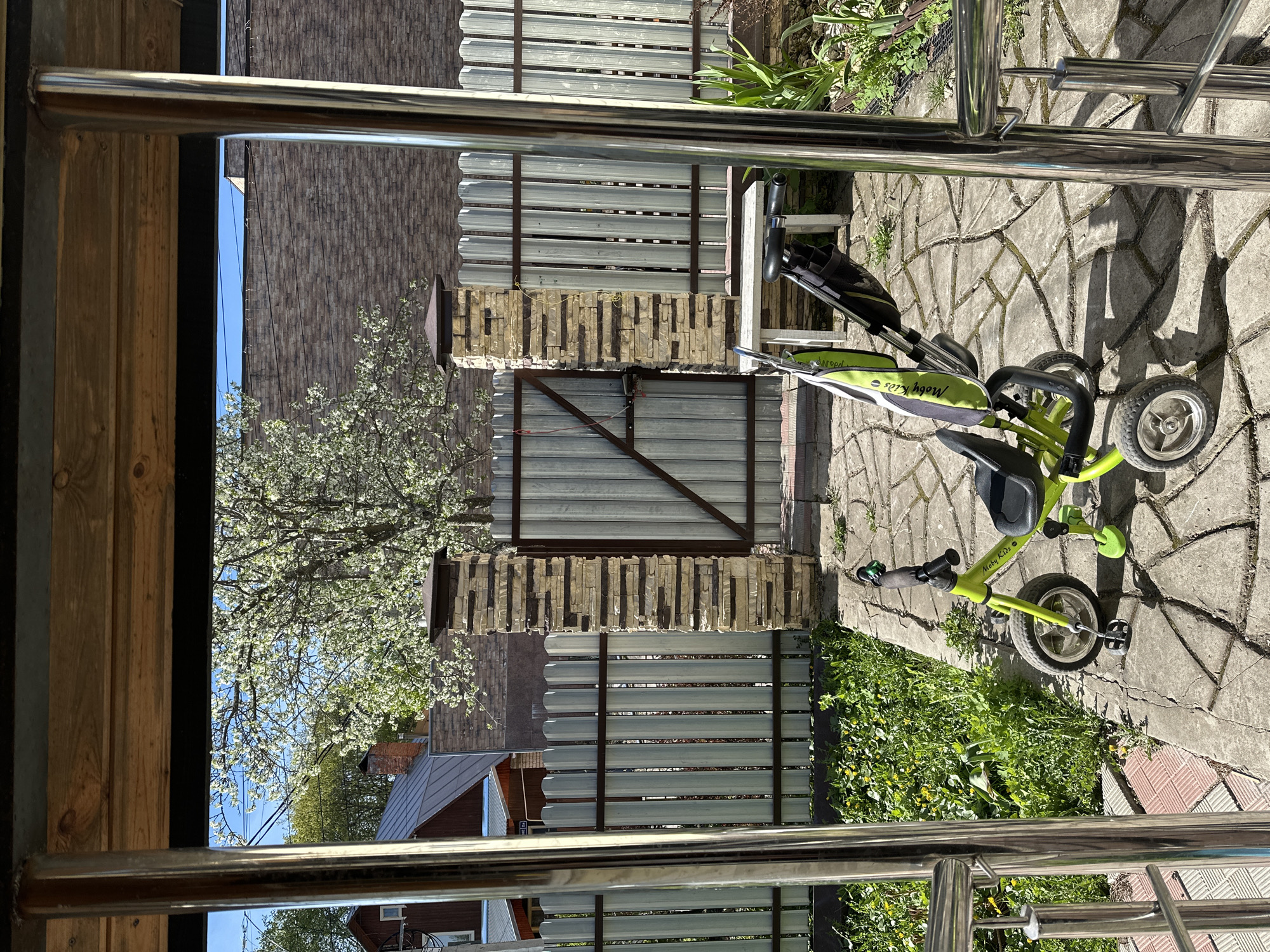 The image shows a view through a partially obscured frame, likely from a balcony or porch, overlooking a backyard or outdoor area in Nizhny Novgorod, Russia. A green children's tricycle ("Moby Kids" brand) is parked on a paved surface, which appears to be a patio or walkway made of stone or concrete slabs. The surface is cracked and covered in some areas by green vegetation. A fence constructed of corrugated metal sheets and stone or brick pillars surrounds the area. Beyond the fence, there are glimpses of houses with varied roofs, suggesting a residential neighborhood. A tree with white blossoms is visible in the distance. The frame through which the scene is viewed is made of wood and stainless steel.