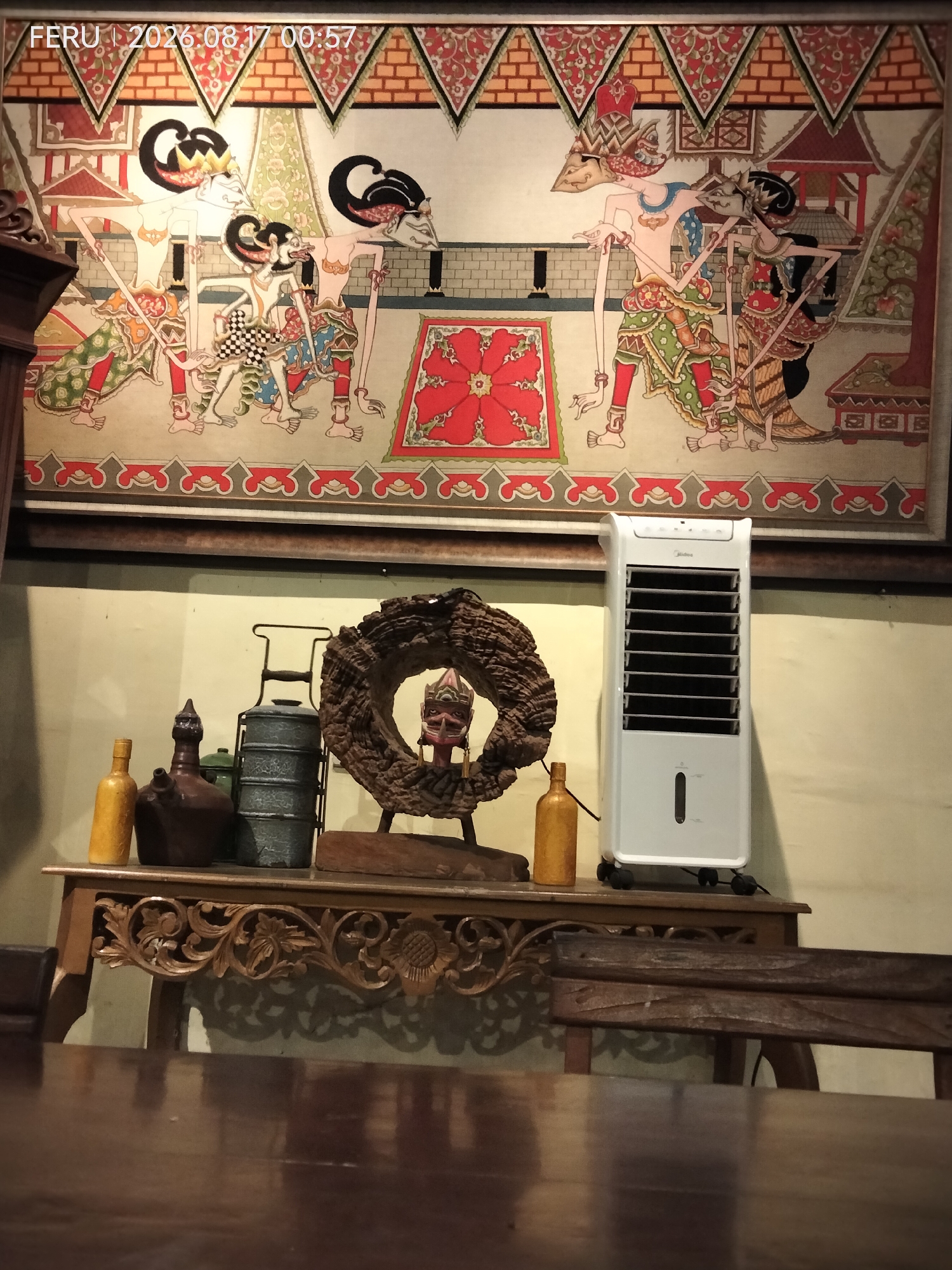The image is a medium shot taken indoors, showing a decorative scene set against a backdrop of a large mural. The mural depicts traditional Indonesian wayang figures in colorful attire, set within an architectural context. Below the mural, a light-colored wall serves as the background for a collection of objects displayed on a wooden table.

On the table, from left to right, are: a yellow glass bottle, a dark brown ceramic jug with a spout, a metallic tiered lunchbox, and a rustic, circular wooden sculpture with a carved mask-like face within its opening. To the right of the objects is a white portable air cooler. The table itself is ornately carved with floral patterns. In the foreground, a dark wooden table with a glossy finish is visible, reflecting the objects on the table above. A portion of a dark wooden chair is also in the foreground. The lighting appears to be warm and somewhat dim, creating shadows.

The text overlay at the top left of the image reads "FERU | 2026.08.17 00:57".

The scene appears to be a domestic interior, possibly a living room or a display area within a home or a shop. The specific location in Sukodono, Indonesia, is inferred from the provided context and is not visually discernible from the image itself.