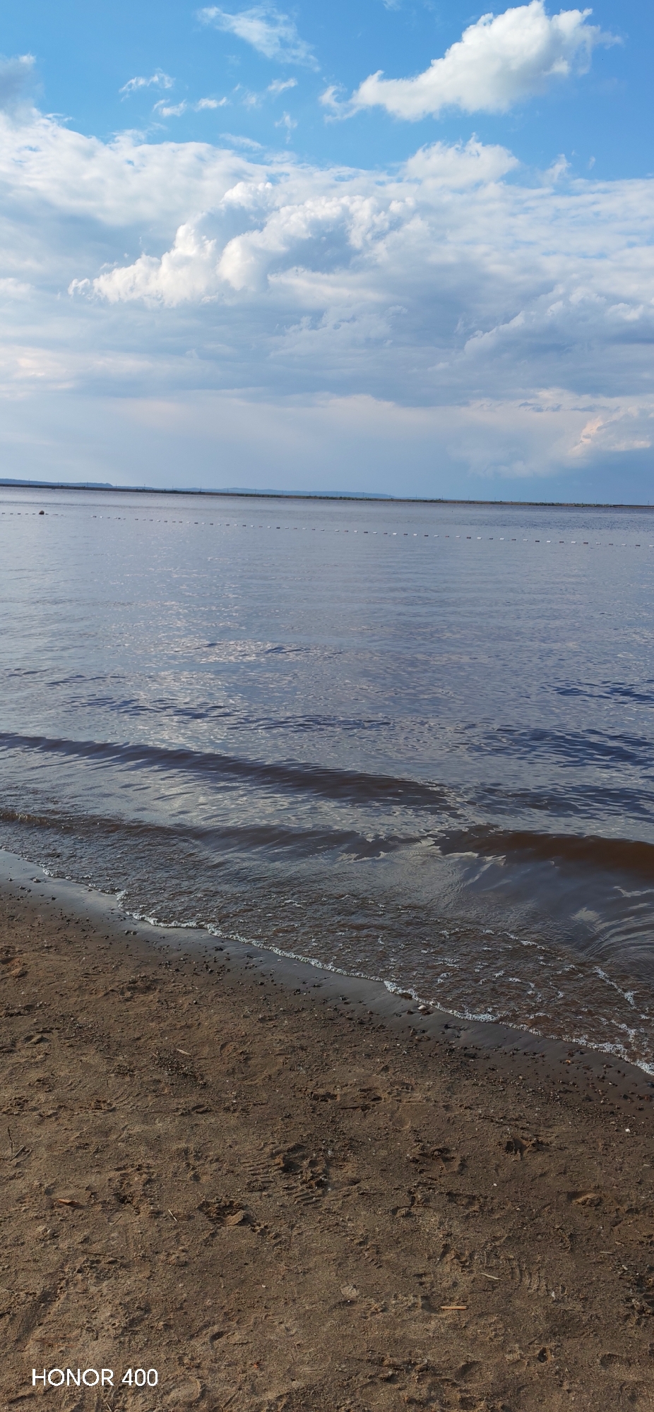 The image depicts a serene view of a body of water, likely a river or a lake, under a partly cloudy sky. The foreground is a sandy beach with some pebbles and debris scattered on it, showing clear indentations that suggest footprints. Gentle waves lap at the shore, creating a foamy edge where the water meets the sand. In the distance, a faint, long line of buoys stretches across the water, possibly marking a boundary or a swimming area. The horizon is visible as a thin line separating the water from a distant, hazy shoreline, which is marked by what appears to be a gentle, rolling landscape under the clouds. The sky is a mix of bright blue patches and soft, gray and white clouds, indicating it might be mid-afternoon with changeable weather. The overall mood is peaceful and tranquil, evoking a sense of calm and the natural beauty of the waterfront. The text "HONOR 400" is visible in the lower left corner of the image, indicating the device used for capture. This scene is likely located in or near Chaikovsky, Russia, given the provided location context.