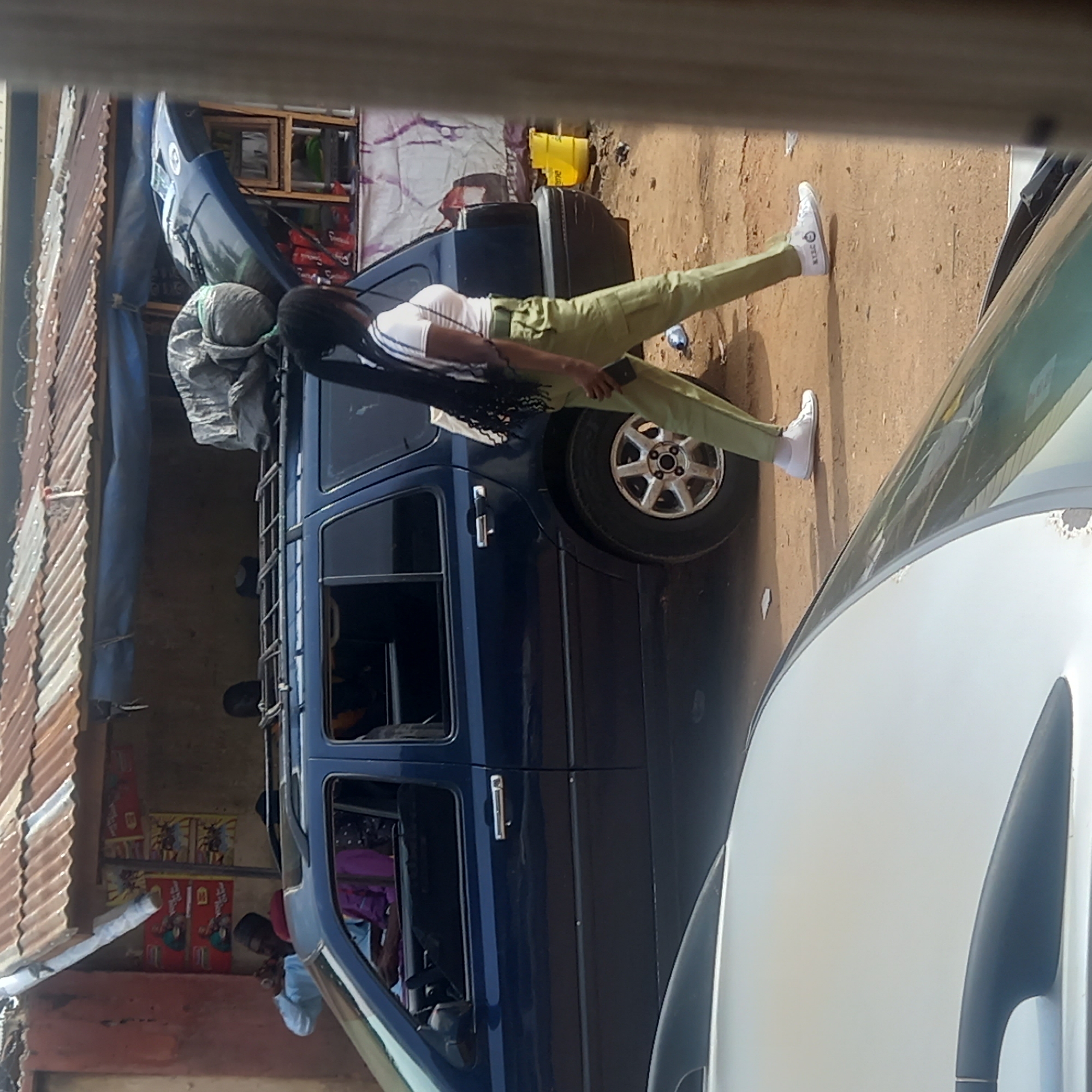 The image shows a woman standing beside a blue van. She is wearing a white top, green pants, and white sneakers with socks. She has long dark hair. The van has luggage strapped to the roof and a ladder on the side. Part of another white vehicle is visible on the right. On the left, there is a building with a corrugated metal roof. The location is Dadi Riba, Nigeria.