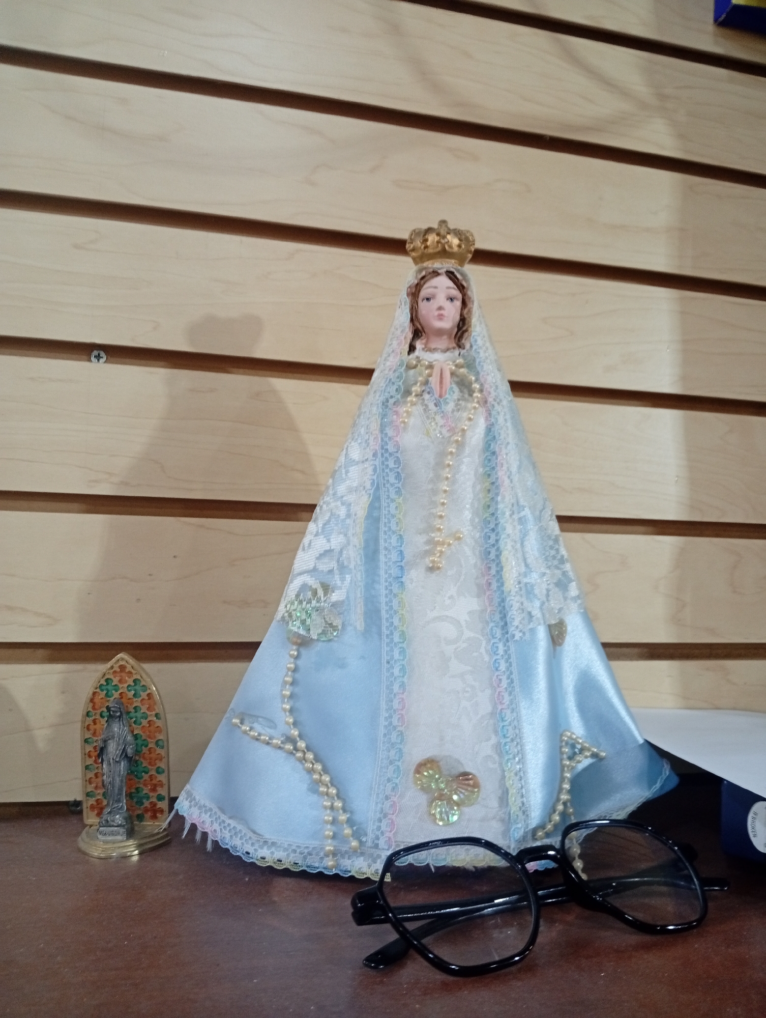 This media file shows a figurine of the Virgin Mary, adorned with a crown and a veil, standing on a wooden surface. The figurine is dressed in a blue and white gown, with a rosary made of pearls draped around its neck and flowing down its front. To the left of the figurine, there is a smaller statue of a religious figure within an arched frame, also with colorful decorative elements. In the foreground, a pair of black-rimmed glasses rests on the wooden surface. The background consists of a light-colored wooden wall with horizontal slats.
The setting appears to be indoors, possibly a home or a place of worship. The lighting is soft, suggesting it might be daytime or an indoor artificial light source. There are no immediate cues about the weather or a specific event taking place. The overall mood is serene and contemplative, associated with religious imagery.
The objects present, namely the Virgin Mary figurine, the smaller religious statue, and the rosary, clearly indicate a religious context. The presence of the glasses might suggest that someone was recently using them or placed them there temporarily. There is no discernible text visible in the image. The location context of Juan Griego, Venezuela, adds a cultural and geographical dimension to the scene, though it doesn't directly influence the visual elements captured.