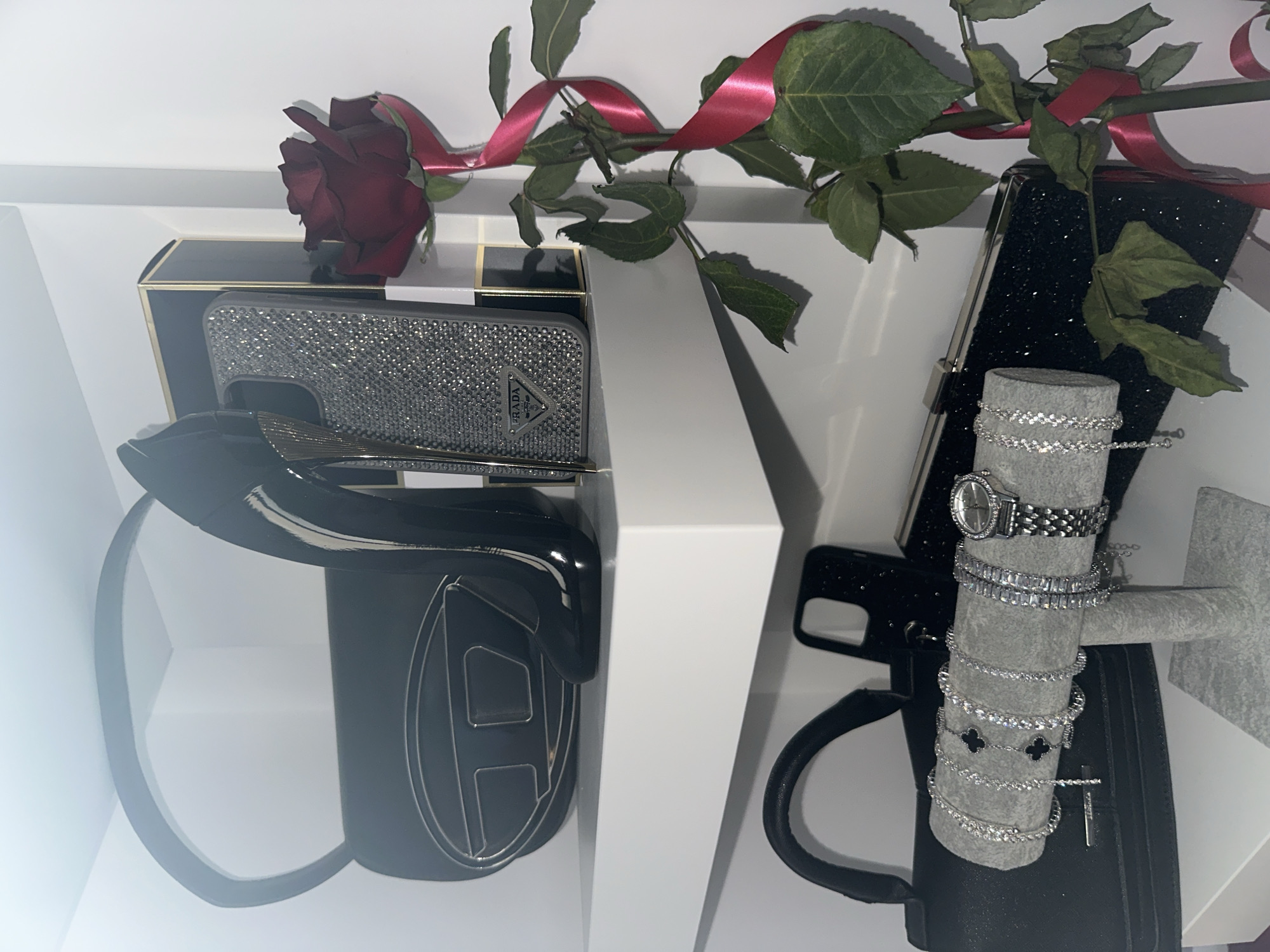The image shows a collection of fashion accessories and a rose arranged within a white, box-like structure, possibly for display or gifting.
On the left side, there is a black purse or bag with a prominent letter "H" design, a bejeweled phone case with a "Prada" logo, and a black box with a gold border. On the right side, there is a black phone case covered in glittering specks, a gray jewelry stand displaying several silver-colored bracelets and a watch. A red rose with green foliage and a red ribbon is draped over the top of the items. The items are presented in a visually appealing manner, suggesting a theme of luxury or gift-giving. The environment is clean and well-lit, typical of a retail display or a studio setting.