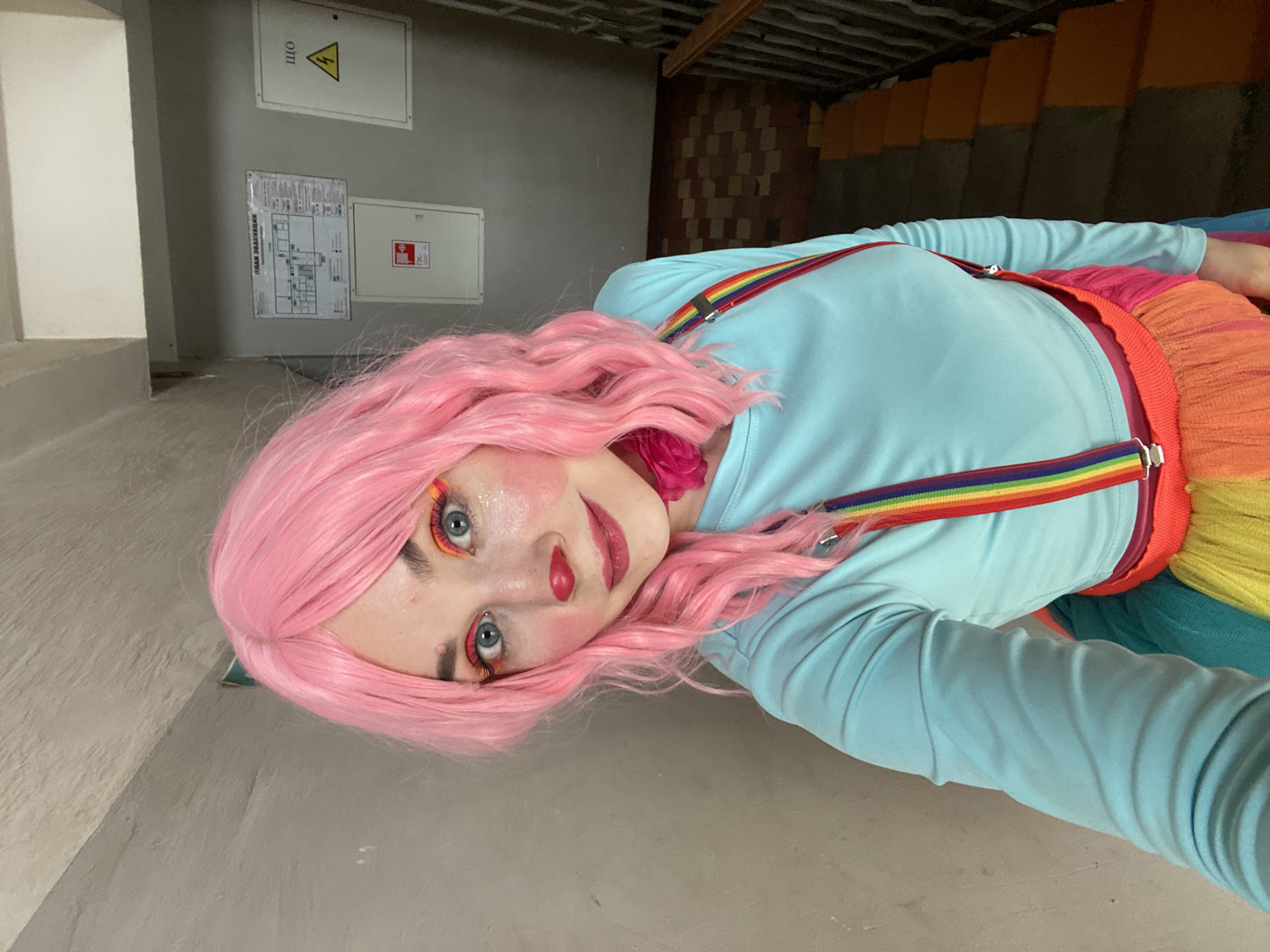 A person wearing a pink wig and clown makeup is taking a selfie in a dimly lit stairwell. The person's face is prominently featured, with bright eye makeup and a red nose and lips. They are wearing a light blue long-sleeved shirt with rainbow suspenders and a colorful tutu. In the background, a stairwell with tiled flooring and a metal railing is visible. A sign with a diagram and Cyrillic text is affixed to the wall, along with a yellow warning sign depicting a lightning bolt. This scene is likely within a residential building in the city of Chaikovsky, Russia, given the architectural style and signage.