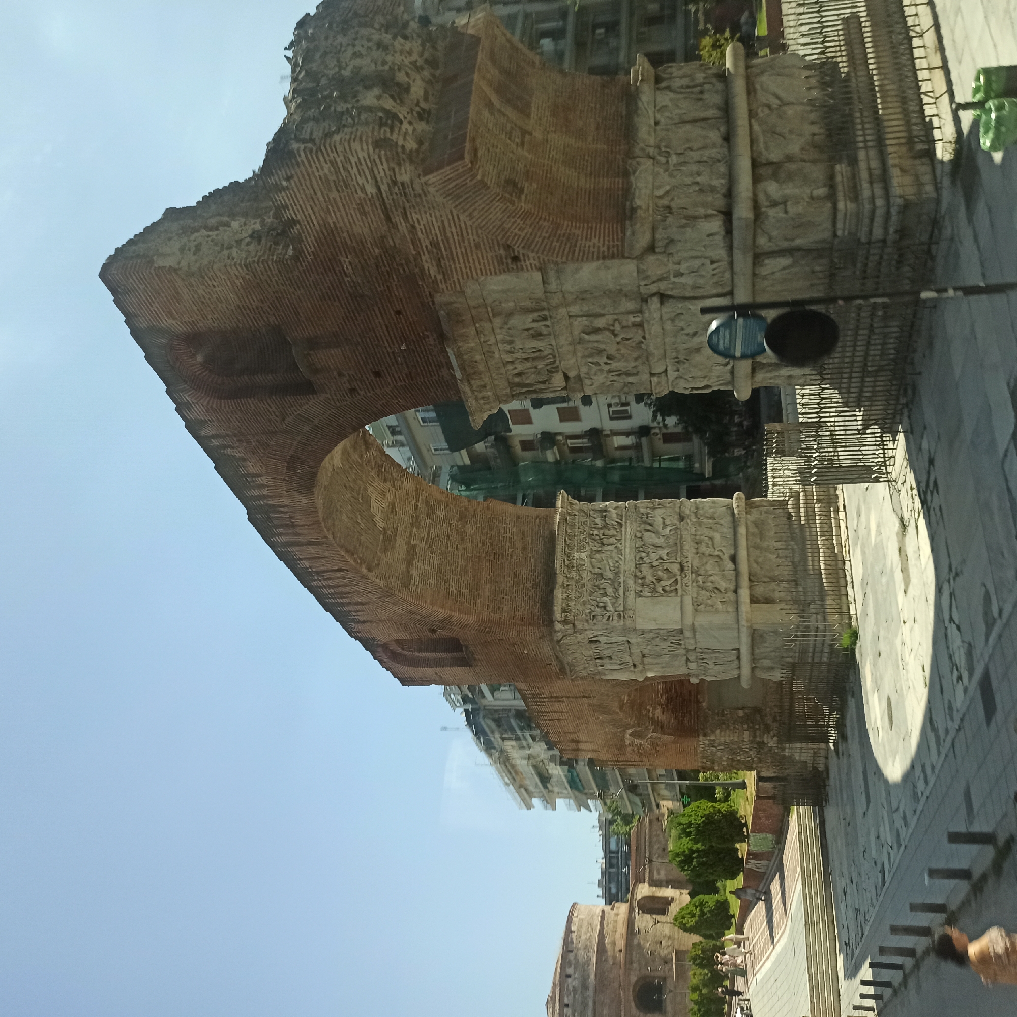 The image shows the Arch of Galerius, a monumental arch in Thessaloniki, Greece. The arch is made of brick and stone, with intricate carvings depicting scenes from Roman mythology and history. It is situated in a public space with a cobblestone pavement and a metal fence surrounding its base. In the background, there are modern buildings, trees, and a clear blue sky. Several people are visible in the distance, walking on a path. To the left of the arch, a circular building, likely the Rotunda of Galerius, is partially visible. The scene appears to be from a sunny day, casting shadows on the ground.
