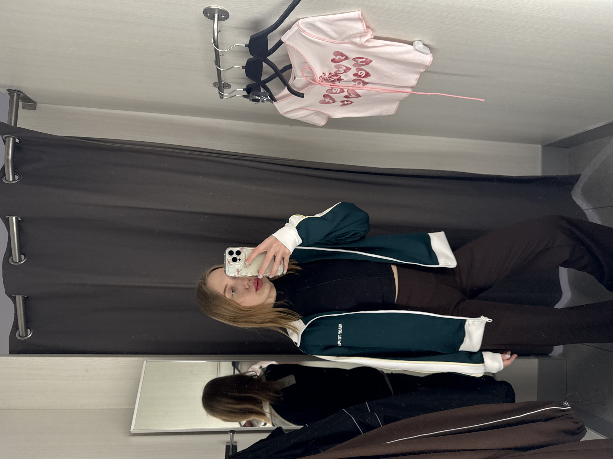 A young adult female is depicted standing in a fitting room, taking a mirror selfie in the city of Ovoshchnoy, Russia. She has light brown hair, a light complexion, and a visible nose piercing. Her attire consists of a dark green track jacket with white and yellow stripes on the sleeves and "READY TO PLAY" text on the left chest, a black high-neck crop top, and dark brown wide-leg pants. She holds a white smartphone with multiple rear cameras in her right hand, angled towards the mirror for the photograph. The fitting room features light gray textured walls, a dark gray curtain on a silver rod, and a dark gray floor. A narrow rectangular mirror on the left reflects the subject's back and a brown garment hanging nearby. On the right wall, a silver metal rail holds three black clothes hangers, one of which displays a light pink short-sleeved crop top adorned with red heart patterns and pink lacing.