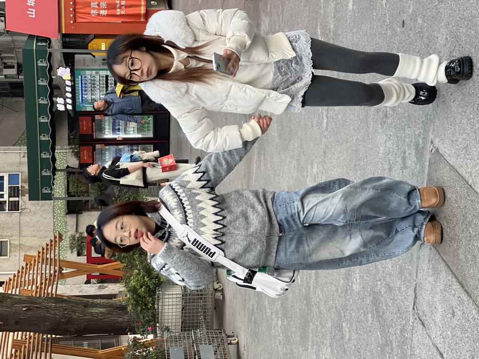 Two young adult females walk forward, holding hands, on a paved outdoor surface in Yuzhong District, China. The female on the left wears a grey zip-up cardigan with a white and dark grey geometric pattern, loose-fitting light-wash blue denim jeans, and tan clogs. She carries a white cross-body bag with a black "PUMA" logo on the strap and wears rectangular glasses; her right hand is near her mouth. The female on the right wears a white puffer jacket with a white faux fur collar, a white long-sleeved top with a white lace-trimmed overlay, dark grey leggings, white ribbed leg warmers, and black patent leather loafers. She holds a silver smartphone in her right hand and wears large, round, dark-rimmed glasses.
In the mid-ground, behind the two women, several individuals are present. A man in a black turtleneck and light pants holds a white bag and a red gift bag, looking upwards. Another man in a blue jacket stands near a row of vending machines and beverage refrigerators, also looking upwards. A green awning extends over a storefront in the background, and red signage with Chinese characters is partially visible on the right. To the left, a large tree with bare branches stands beside a wooden trellis structure and some green bushes in planters. The ground is grey paving.