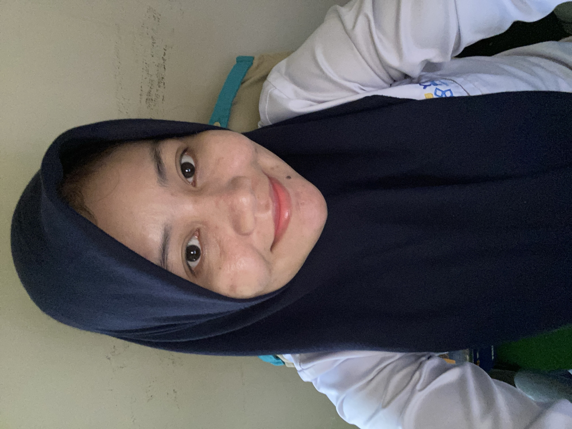 A selfie taken by a woman wearing a dark blue hijab and a white collared shirt. She is smiling and looking at the camera. The background appears to be an indoor setting with a textured wall. There is a hint of a blue and yellow logo on her shirt. The image was likely taken in Banjarbaru, Indonesia, based on the inferred location.
