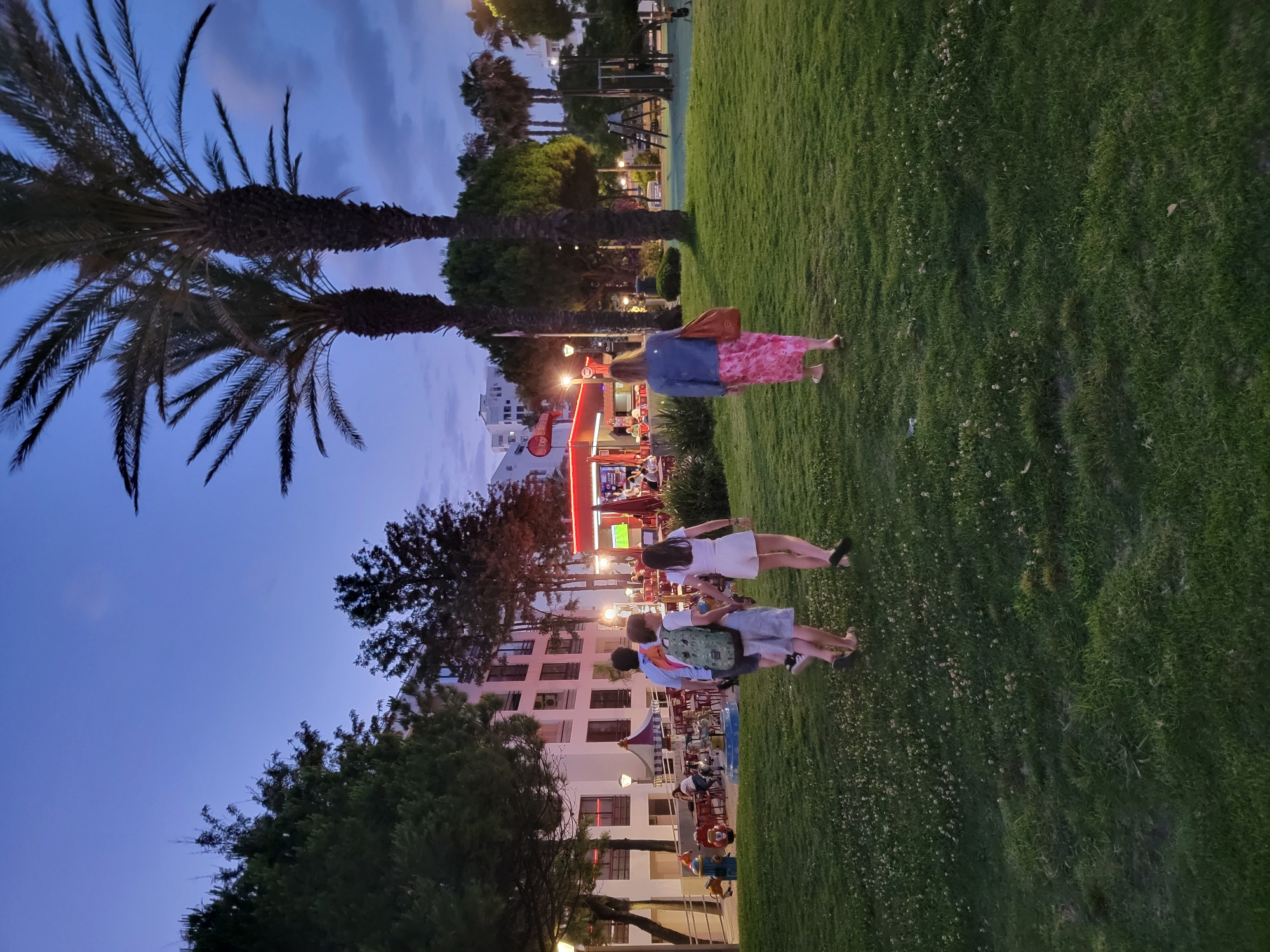 The image depicts an outdoor scene in Quarteira, Portugal, during twilight. In the foreground, a group of four individuals is walking across a well-maintained grassy area dotted with small white flowers. The group consists of three children and one adult. The child in the front, facing away from the camera, wears a backpack. The child behind them wears a white t-shirt and shorts. The third child is dressed in a white t-shirt and white shorts. The adult, also facing away, is wearing a denim jacket and a long pink floral dress, carrying a brown tote bag.
Further in the background, there are palm trees and other trees. To the left, a multi-story building with numerous windows is visible, with lights on inside some of them. Adjacent to the building is an area with red tables and chairs, suggesting an outdoor dining space. A small carousel with animal figures is also present in this area. Further to the right, a red structure, possibly a food stall or shop, is illuminated. A playground with swings and other equipment is also visible on the right side of the image. Streetlights are scattered throughout the scene, casting a warm glow. The sky is a deep blue, characteristic of dusk. The overall atmosphere suggests a public space, possibly a park or promenade, with leisure and dining facilities.