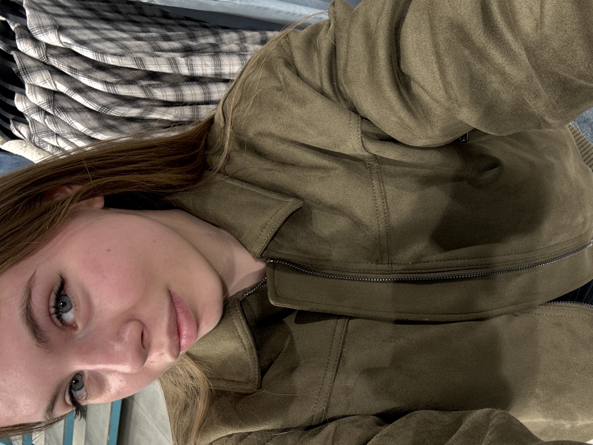 This image depicts a medium close-up, vertical photograph of a young Caucasian woman, approximately late teens to early twenties, situated within a commercial retail clothing environment. The scene is located in the city of Sochi, Russia.
The woman occupies the majority of the frame, positioned slightly off-center to the left. She has light skin, prominent blue eyes with dark eyelashes, light brown eyebrows, a small nose, and full, slightly parted pink lips. Her long, straight light brown hair frames her face and extends over her shoulders. She wears an olive-green, suede-like jacket featuring a collar, a visible silver-colored zipper down the front, and distinct stitching details on the collar and shoulders. A small portion of light blue fabric, likely denim, is visible beneath the jacket on the lower left. Her gaze is directed slightly to the left of the camera, indicating a self-portrait or a posed photograph.
The background is blurred, consistent with a shallow depth of field. To the left, indistinct light blue horizontal stripes or elements are visible. To the right, a rack of multiple plaid button-up shirts, primarily in white, gray, and blue patterns, are clearly visible hanging on hangers, reinforcing the retail store setting. The lighting is even, consistent with indoor artificial illumination. There are no other individuals or active interactions visible within the frame.