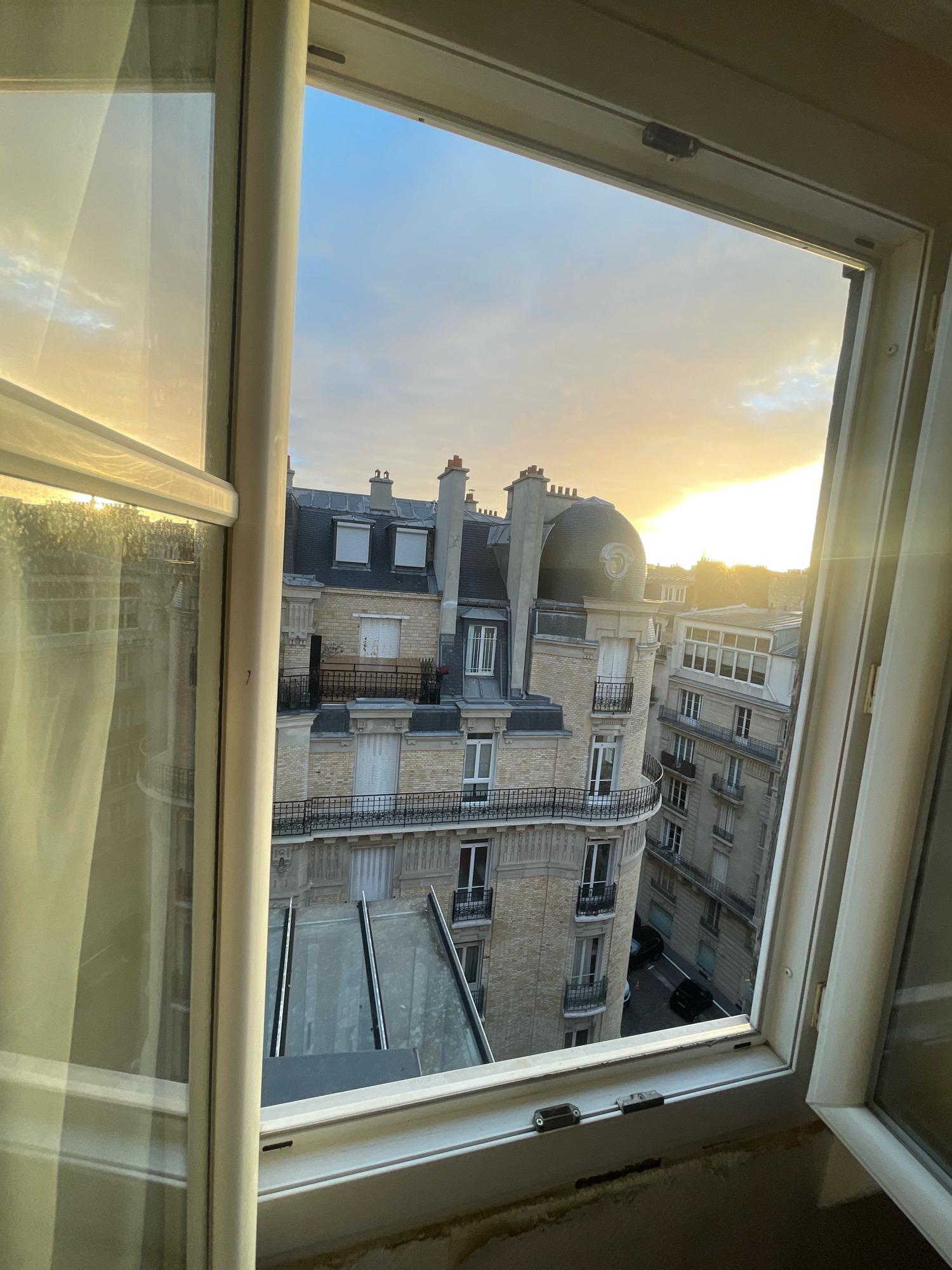 The image shows a view from an open window looking out at Parisian rooftops at sunset. The sky is filled with golden light, and clouds are visible. The architecture is typical of Paris, with ornate buildings, mansard roofs, and chimneys. In the foreground, the white window frame is visible, as is a portion of a sheer curtain. The view looks down onto a street with parked cars, suggesting an urban setting.
