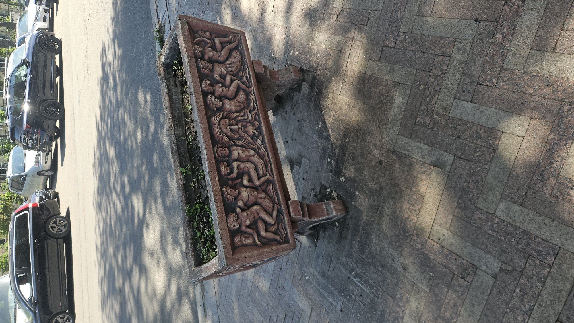 The image shows an ornate rectangular planter box sitting on a paved sidewalk near a street. The planter box is decorated with bas-relief figures, seemingly human forms in various reclining poses. Small green plants are growing inside the planter. The sidewalk is made of rectangular bricks in a herringbone pattern. A row of parked cars is visible on the left of the street and the sidewalk, with shadows cast across the pavement and vehicles. Based on the presence of the planter and architectural style, it is possible that this image was taken in Chișinău, Moldova.