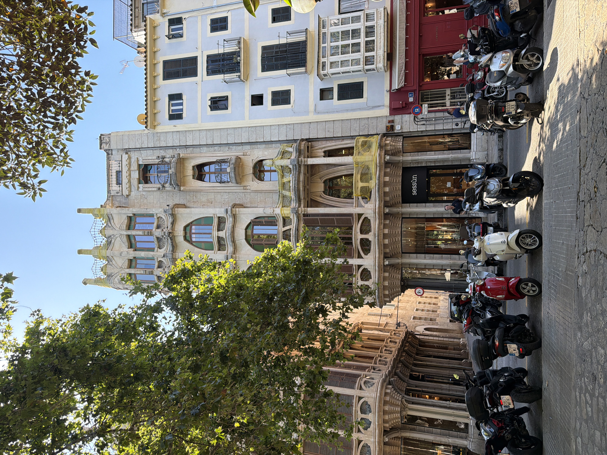 This image captures a street scene in Palma, Spain, with a prominent building in the center displaying ornate architecture, featuring arched windows, balconies, and decorative stonework. To the right, a lighter blue building with balconies and shutters stands next to it. In front of the buildings, a row of parked motorcycles and scooters occupies the paved street. A large tree with green leaves partially obscures the view of the buildings, casting shadows on the scene. The sky is clear and blue. People are visible on the sidewalk, walking or standing near the buildings. One person is standing in the doorway of a shop with the name "sessùn" visible. The overall impression is a sunny day in an urban environment with historical architecture.