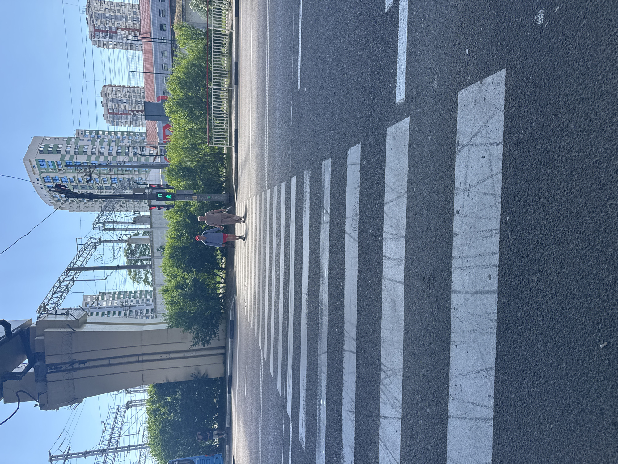 Two people are walking across a pedestrian crossing on a sunny day in Sochi, Russia. The scene is set in an urban environment with tall buildings and power lines visible in the background. The pedestrian crossing is clearly marked with white stripes on the asphalt road. The sky is clear and blue, indicating good weather conditions. The time of day appears to be daytime, likely late morning or early afternoon, given the angle of the sun and the shadows. The people are casually dressed, and their expressions are not discernible from the image. One person is wearing a light-colored shirt and dark pants, while the other is wearing a red hat and shorts. The overall atmosphere is neutral and everyday.