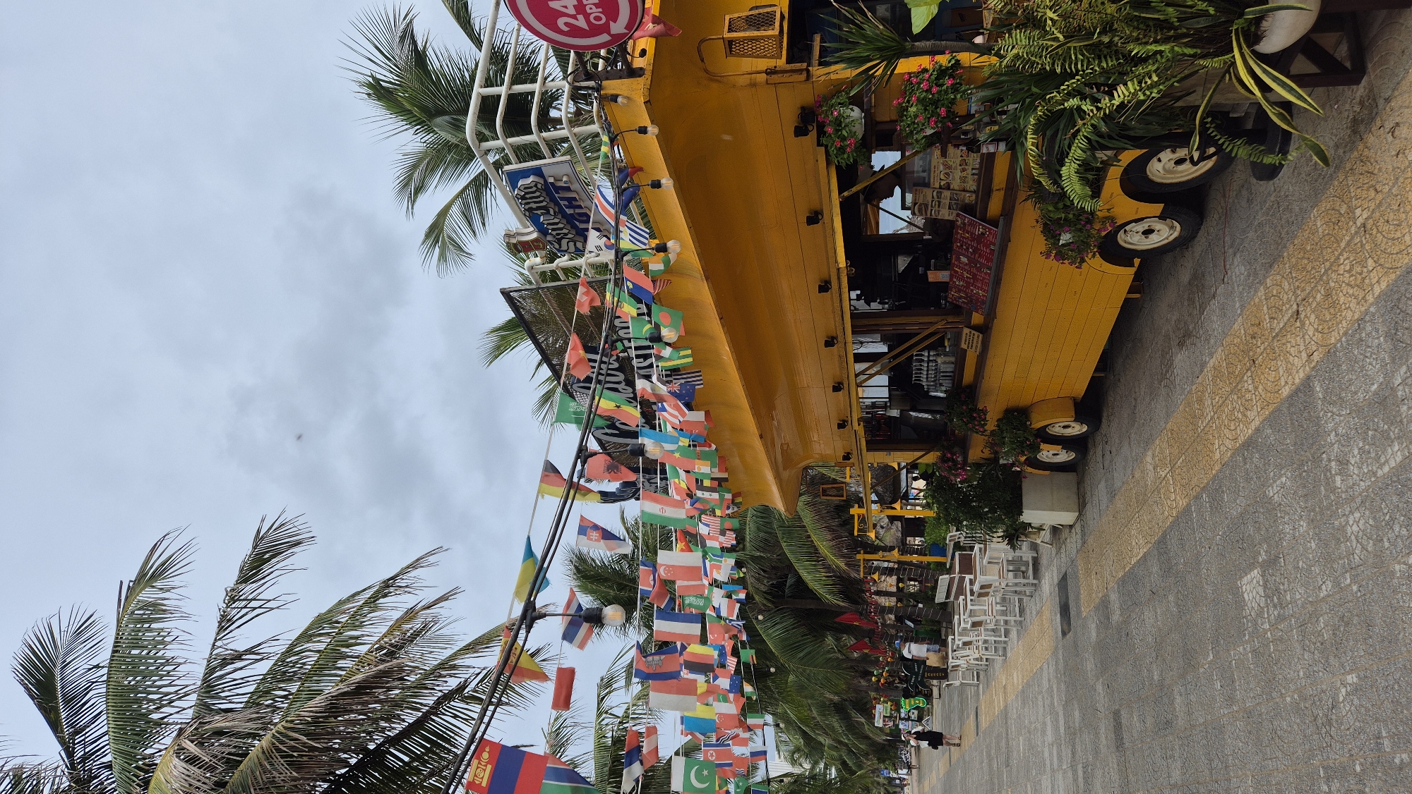 A bright yellow food truck, designed to resemble a bus, is parked on a paved street lined with palm trees and decorated with numerous small international flags strung overhead. The food truck has an open serving window displaying menus and beverages. White tables and chairs are set up on the sidewalk in front of the truck, suggesting an outdoor dining area. Several people are visible walking along the sidewalk in the background. The sky is overcast, and the overall atmosphere suggests a commercial area, possibly a beachside promenade in Da Nang, Vietnam, given the tropical foliage and pedestrian activity.
