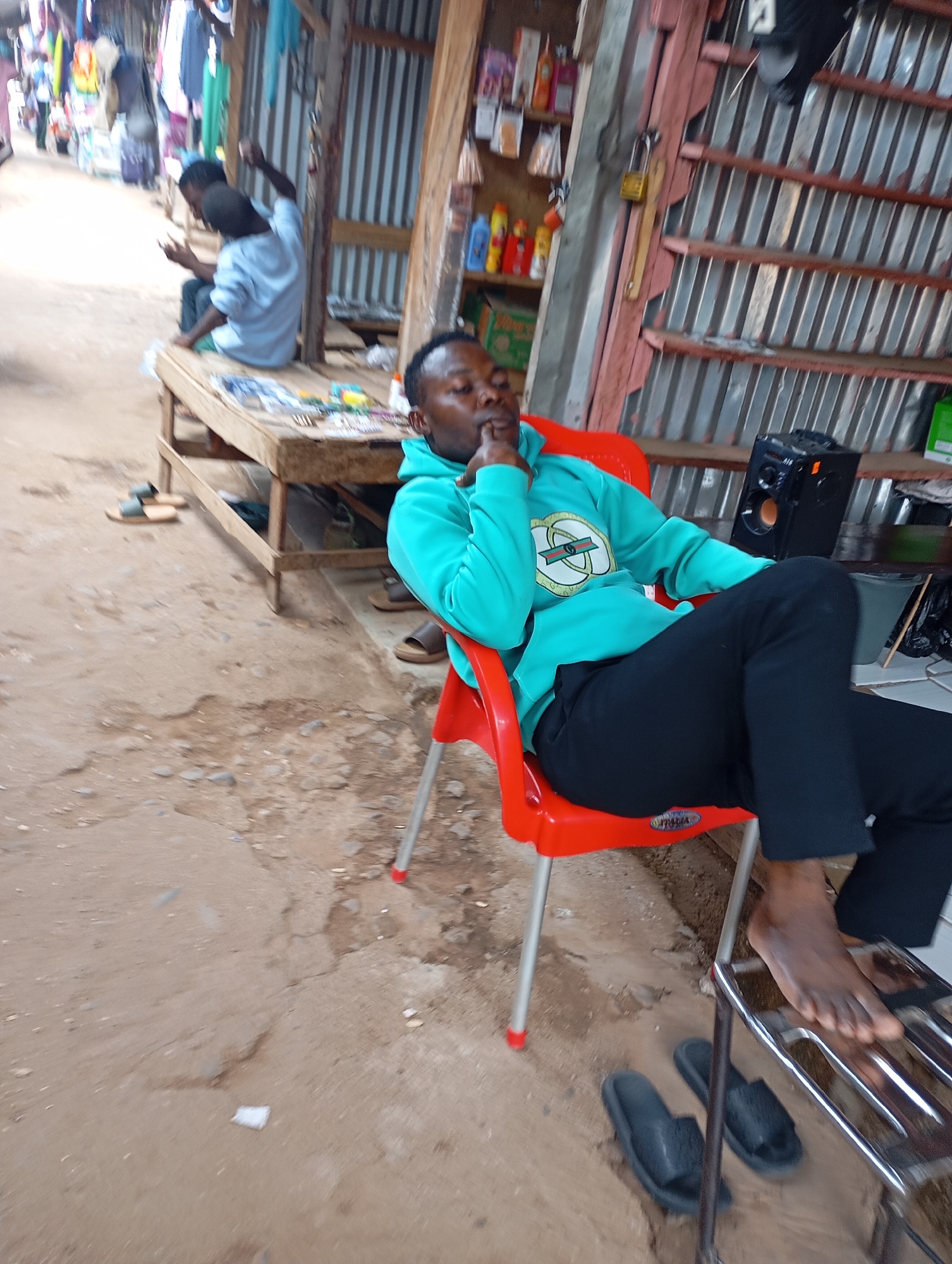 A young man in a teal hoodie and black pants is seated on a red plastic chair in an outdoor market setting. He is looking down and resting his chin on his hand. To his right is a metal shelf with a black speaker on it. To the left of the chair are a pair of black slippers, and to the right of the chair, partially visible, is another pair of brown slippers. In the background, a wooden table is covered with various items, and behind that, a corrugated metal structure appears to be a stall or shop. Further in the background, another person in a light blue shirt is seated. The ground is a mixture of dirt and concrete. The scene is consistent with an open-air market in Shendam, Nigeria, likely in the morning or afternoon given the lighting.
