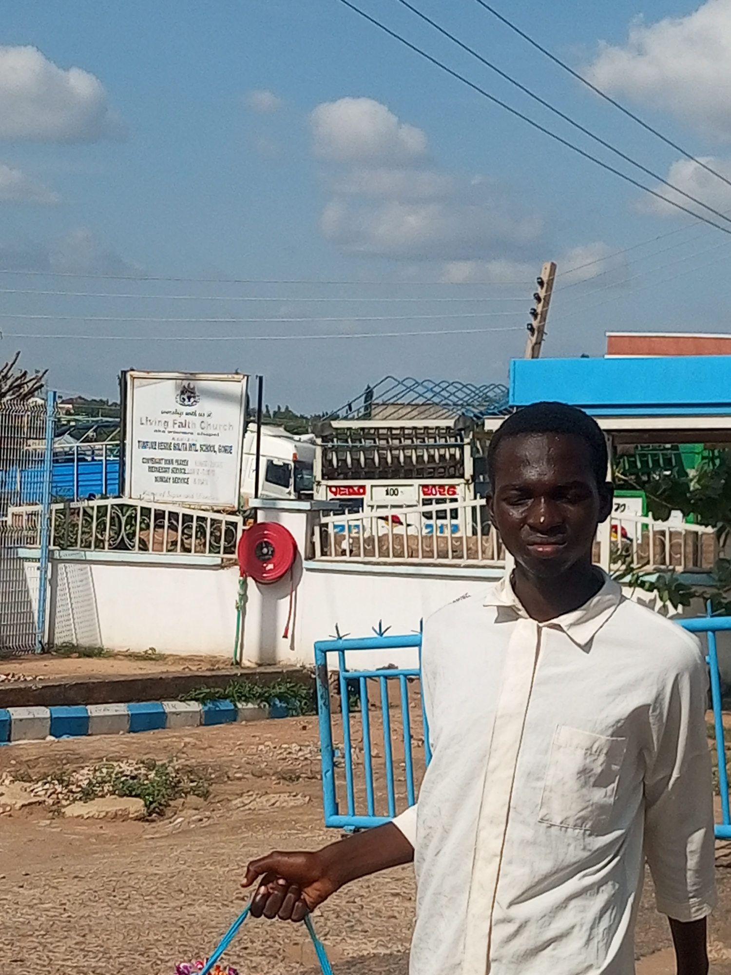 A young Black male, wearing a light-colored button-down shirt, stands in front of a white building with a sign that reads "Living Faith Church" in English. Behind the man, a portion of a blue structure with red and orange accents is visible, possibly a gas station. The background includes a clear sky with scattered clouds, power lines, and a fence. A red fire hose reel is mounted on the wall of the white building. The ground appears to be a mix of gravel and dirt, with a blue and white striped curb visible in the foreground. A blue metal barrier is also present. The overall scene suggests an outdoor setting in a town or city, possibly Garko, Nigeria, given the church's name and the presence of a gas station.