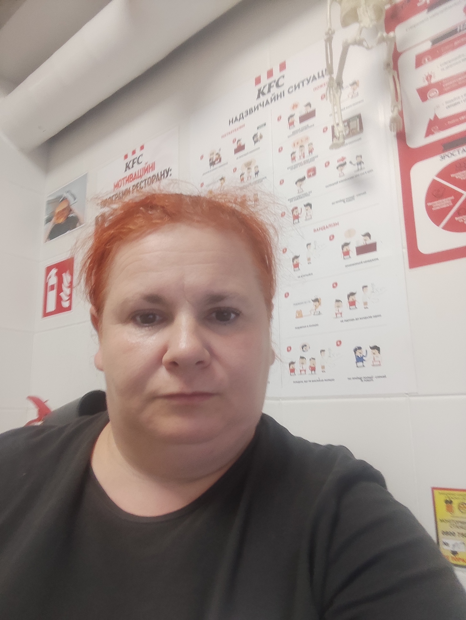 A woman with short, reddish-orange hair is taking a selfie indoors. She is wearing a dark t-shirt. Behind her, on a white tiled wall, are several posters with red and white graphics and text in Ukrainian. One poster has "KFC" and "НАДЗВИЧАЙНІ СИТУАЦІЇ" (emergency situations) at the top, with a series of illustrations below depicting different scenarios. Another poster has "KFC" and "МОТИВАЦІЙНІ ПРОГРАМИ РЕСТОРАНУ" (restaurant motivational programs). To the left of these posters is a framed image showing a person from the shoulders up. To the left of that, a red fire extinguisher symbol is visible on the wall. A skeletal figure is hanging from the ceiling or a wall fixture in the upper right portion of the image. Another poster with a circular graphic in red and white is partially visible on the right. The overall environment appears to be a commercial establishment, possibly a KFC restaurant in Chernivtsi, Ukraine, given the visible signage and text.