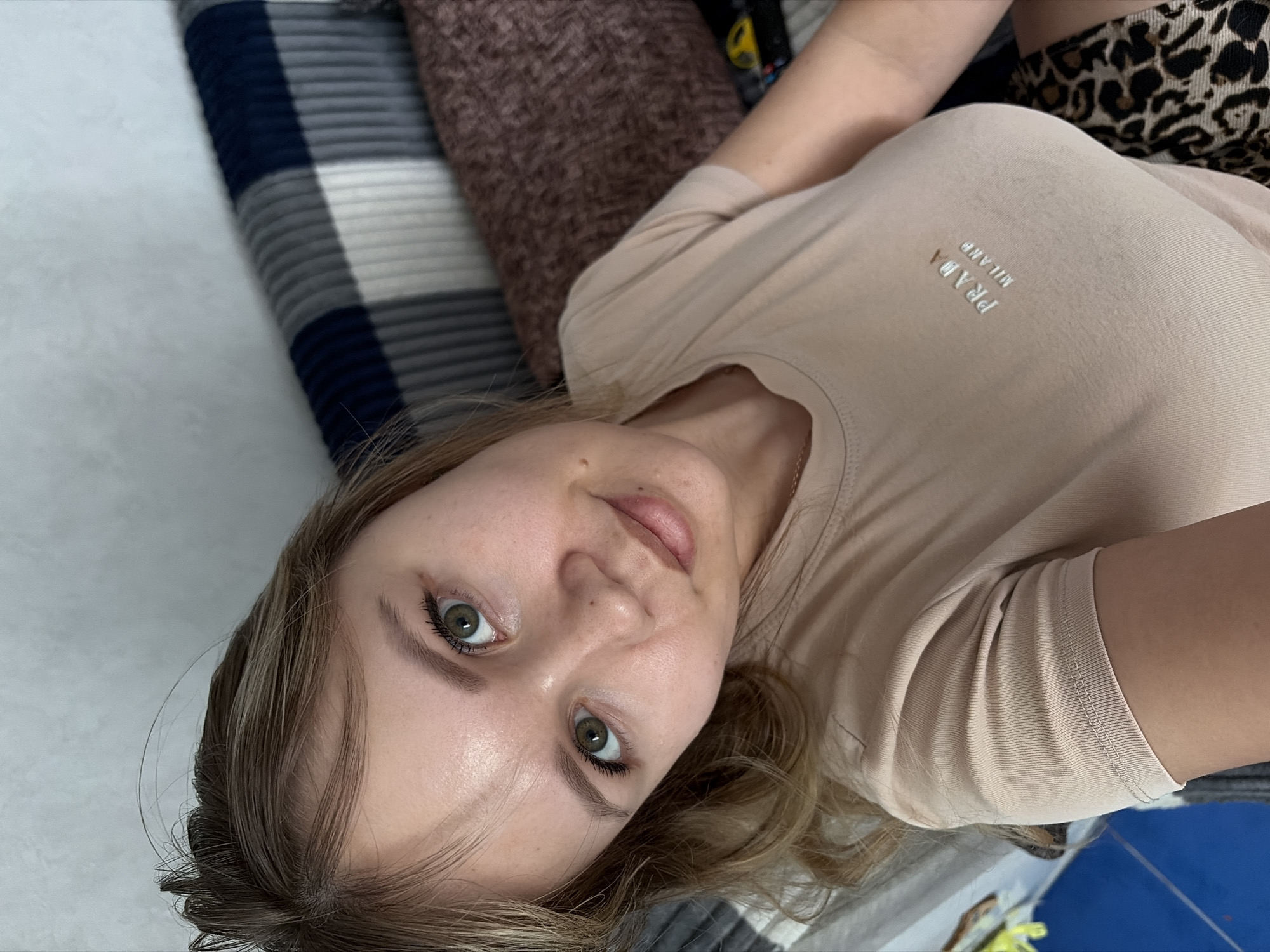 A young woman with light brown hair and prominent green eyes is depicted in a close-up, slightly rotated portrait, likely a selfie. She is lying down, looking directly towards the camera with a neutral to slightly pleasant expression. Her eyelashes have mascara, and her skin appears natural.
She is wearing a light beige short-sleeved t-shirt, on the chest of which the metallic silver text "PRADA MILANO" is visible. A hint of a gold chain can be seen near her neck. Part of her attire includes a leopard print fabric, possibly shorts or a skirt, visible near her right shoulder and arm.
The setting appears to be indoors, suggesting a relaxed home environment such as a bed or sofa. To her left, a striped blanket or sheet with dark blue, grey, and white bands provides a patterned background. To her right, a textured brown cushion or pillow is partially visible. The lighting is bright and even, indicating daytime. The image captures a candid moment in a casual indoor scene.