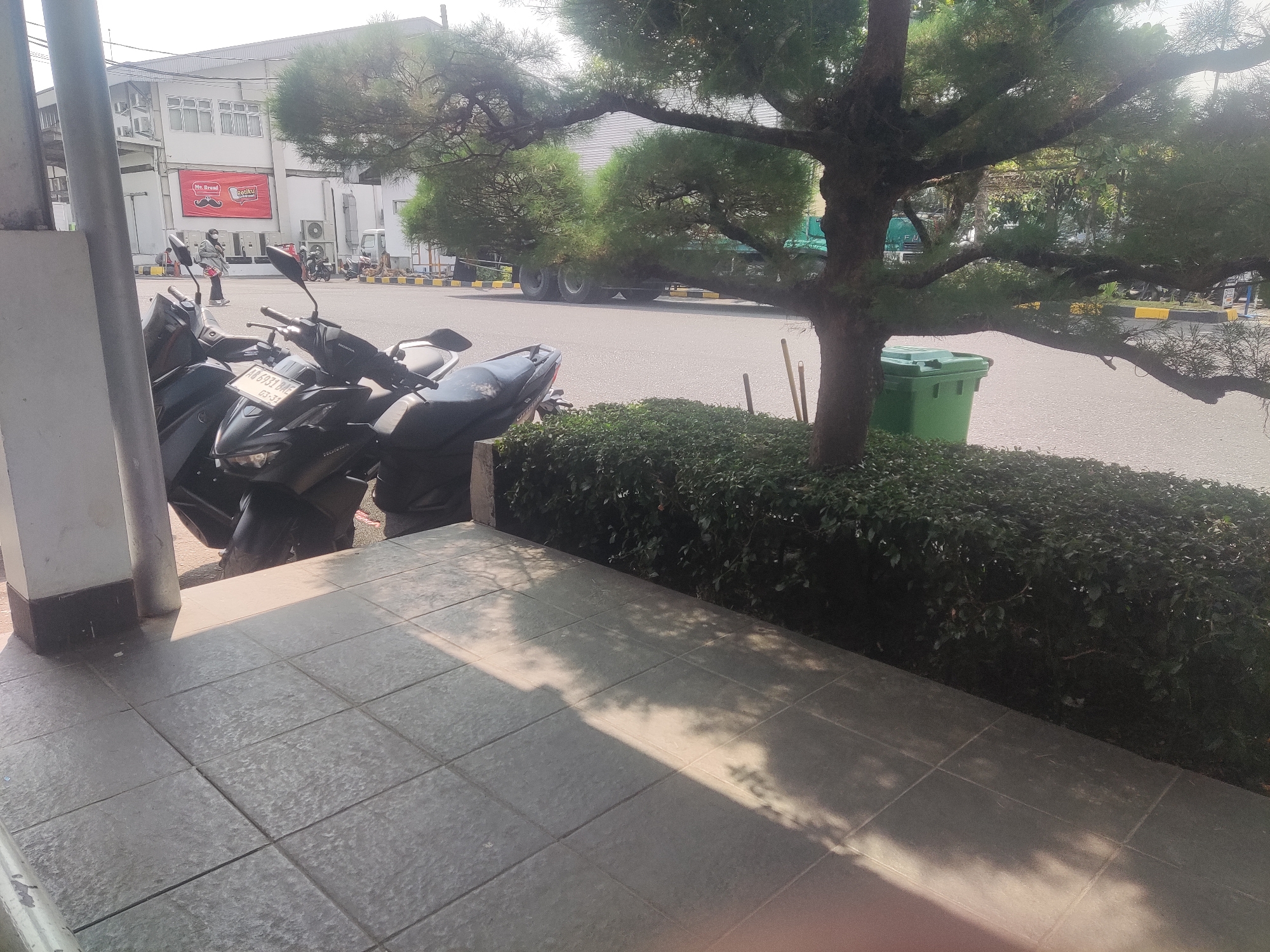 In Trihanggo, Indonesia, a row of parked motorcycles is visible in the foreground. Beyond them, a lush green hedge separates the tiled walkway from a paved road. In the background, a white building with numerous windows is seen, with a large advertisement for "Mr. Bread" displayed prominently on its facade. A person wearing a mask and a jacket stands near the advertisement, seemingly observing something. Further down the road, a large green truck is parked. The scene is illuminated by bright sunlight, casting shadows on the walkway. A green trash bin is situated next to a tree with a sprawling canopy.