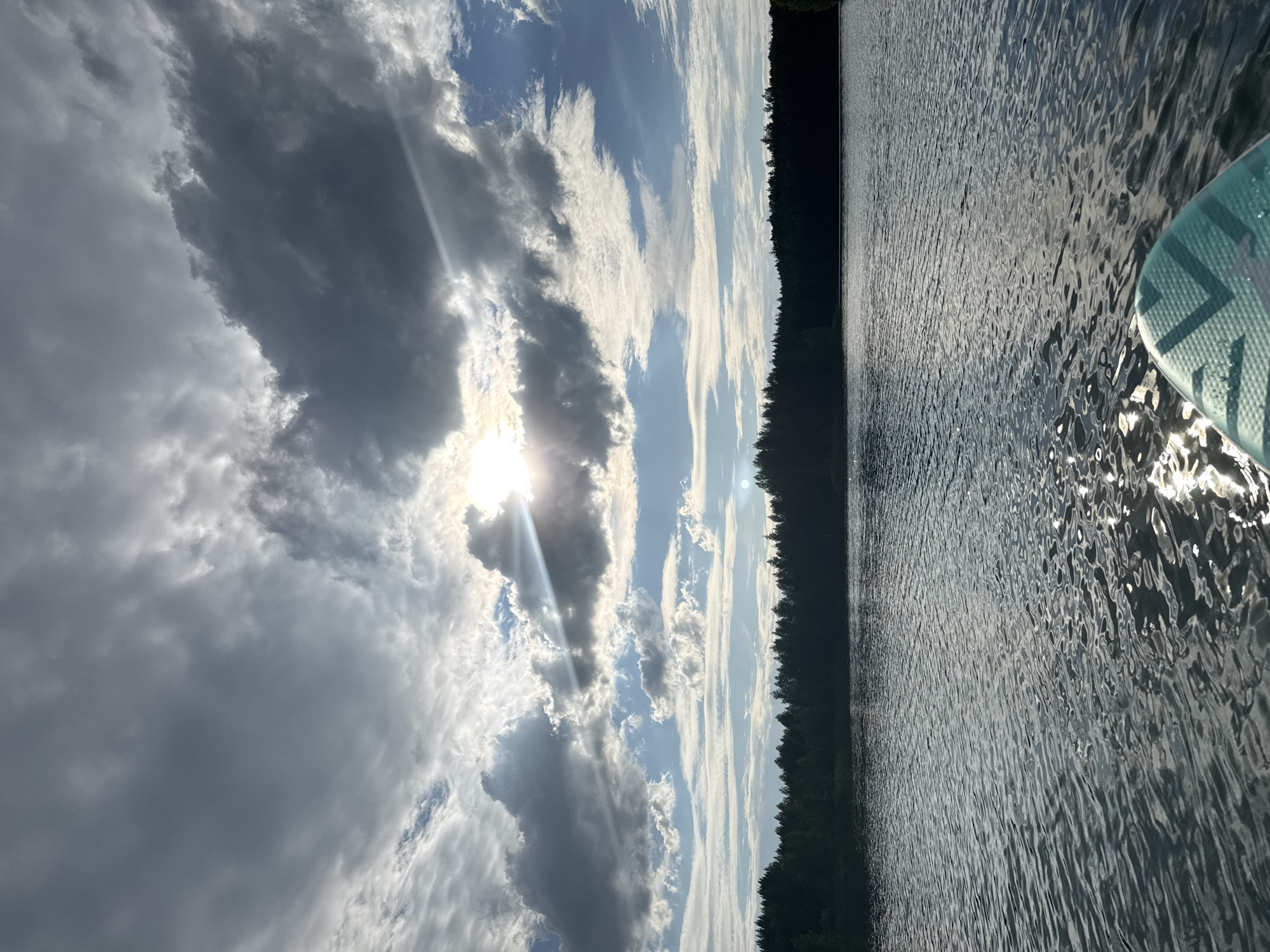A wide shot captures a serene lake scene, presumably in Novyi, Russia, under a dynamic sky. The foreground features the tip of a paddleboard, its turquoise and white patterned surface visible. The lake's surface is rippled, reflecting the sunlight in shimmering patterns. A dark, dense treeline forms the horizon, creating a stark contrast against the sky. The sky itself is a dramatic canvas of clouds, with patches of bright blue sky peeking through. The sun, partially obscured by heavy, dark clouds, casts brilliant rays across the scene, illuminating the clouds and water. The overall atmosphere is one of natural beauty and tranquility.