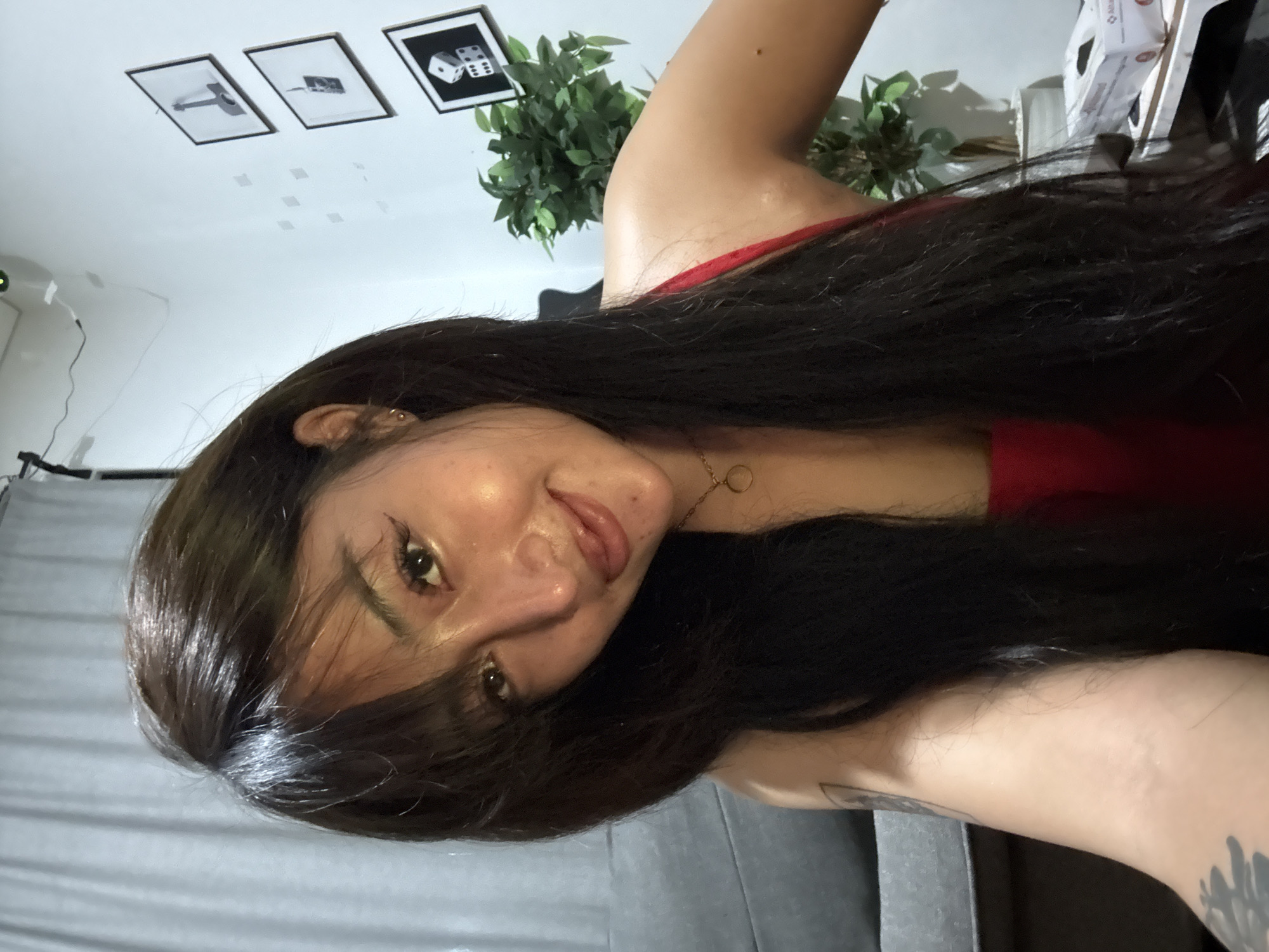 The image displays a headshot and upper body of a young woman, captured with a 90-degree clockwise rotation, making her appear to be lying down with her head on the left side of the frame. She has long, dark, straight hair cascading across her chest and shoulder, partially covering her face. She looks directly at the camera with a gentle smile and a pleasant expression.
The woman has fair skin, dark eyebrows, and wears light eye makeup including prominent eyelashes. A thin golden necklace with a circular pendant adorns her neck, and a small golden hoop earring is visible in her left ear. She is wearing a red top, visible around her shoulder and chest. A dark, possibly abstract or floral, tattoo is visible on her right forearm/bicep.
The setting appears to be indoors. Behind her head and upper body, a light-colored wall or ceiling is visible, featuring three framed pictures. From left to right in the current orientation: one depicting a minimalist illustration of a hammer, another with an unclear subject, and the third showing two dice (one displaying '6', the other '2' or '5'). Small, square, reflective elements are also affixed to this surface. A leafy green plant adds a natural touch behind her right shoulder, and some cardboard boxes are partially visible further in the background. Beneath her, a gray textured surface, possibly a sofa or bedding, is present. The scene is brightly and evenly lit. There is no legible text visible in the image.