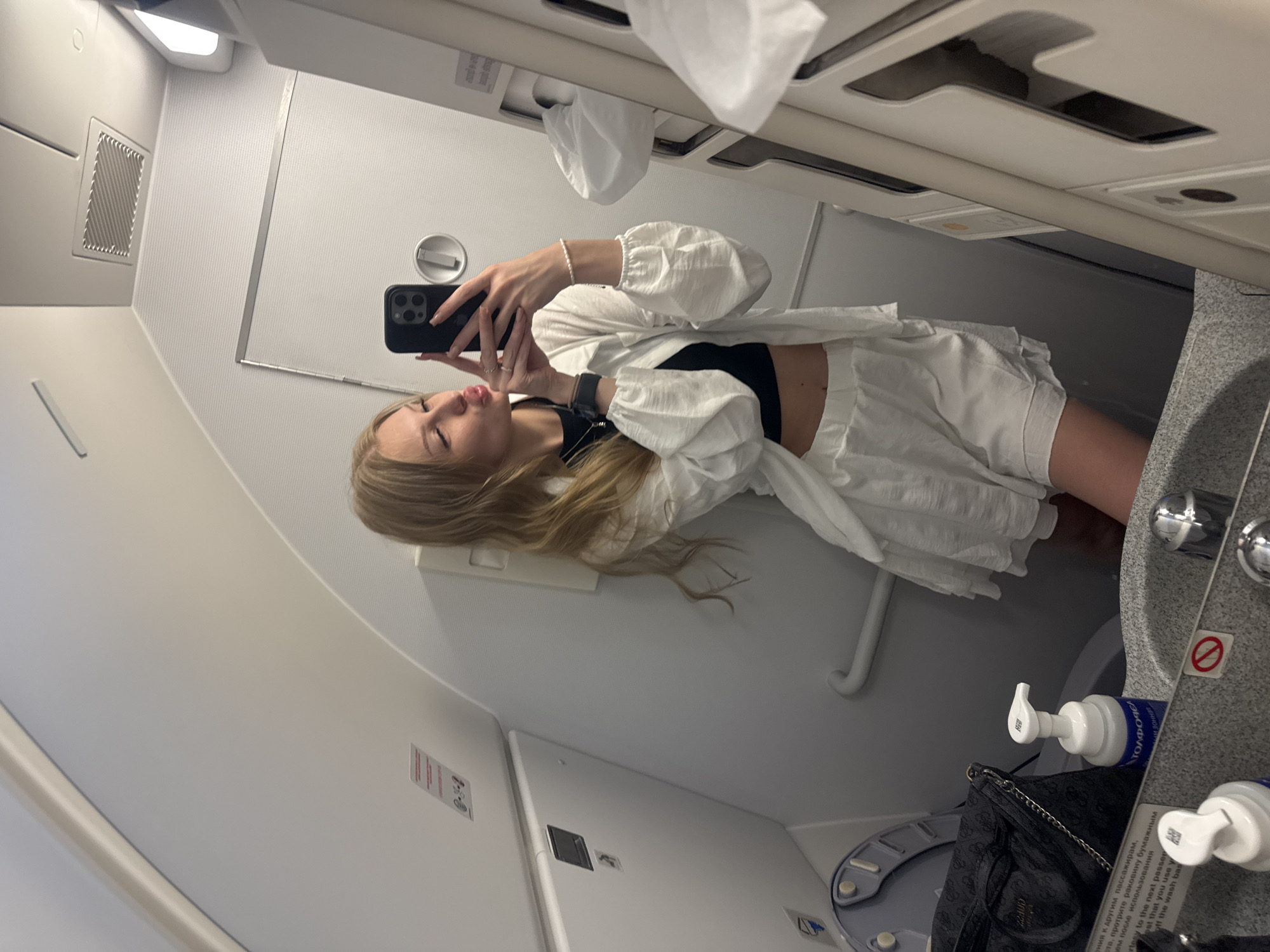 A young blonde woman is taking a selfie in a small airplane lavatory. She is wearing a white two-piece outfit and has her phone in her right hand. The lavatory has a toilet, sink, and a mirror. The walls are white and the lighting is bright. The image does not provide any information about the city of Kemer, Turkey.