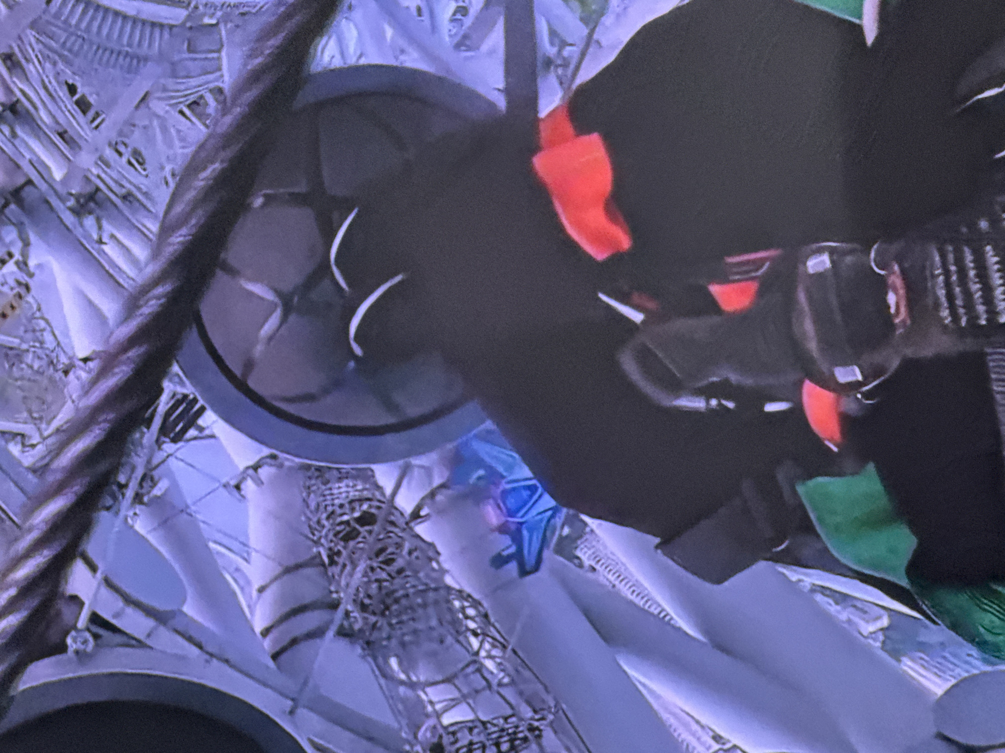 The image shows a person dressed in dark clothing with red and green accents, wearing a helmet or head covering. They are holding onto what appears to be a rope or cable. The background is blurry but suggests an industrial or construction environment with metallic structures and wires. There are no discernible landmarks that place this image in Moscow, Russia.