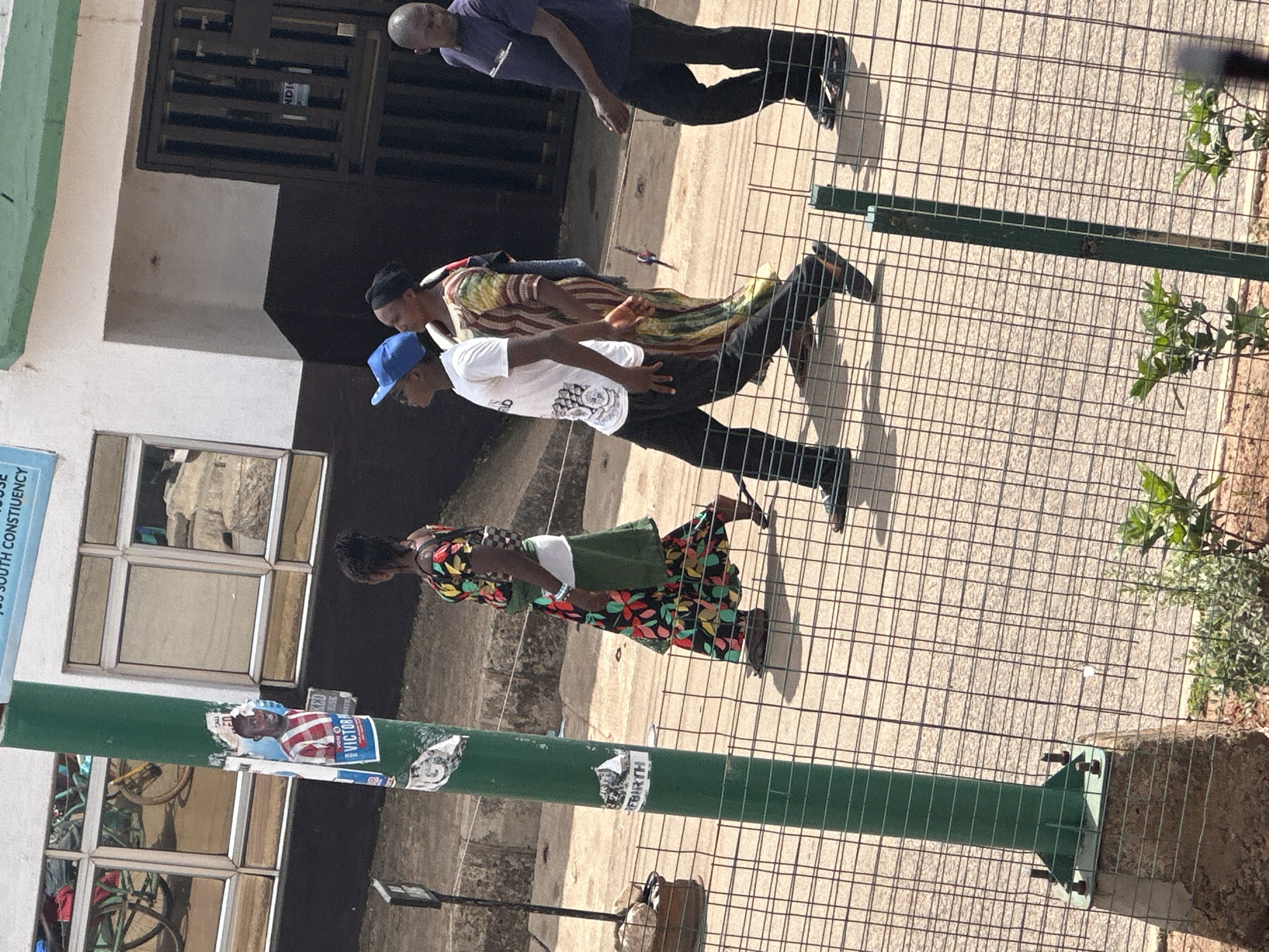 The image depicts a street scene in Bukuru, Nigeria, under bright daylight. A green lamppost stands prominently in the foreground, adorned with posters. Behind the lamppost, a group of four individuals are walking on a paved surface, separated by a wire fence.
The first person, a woman, is on the left, wearing a colorful floral dress and a green sash. She is followed by a young person wearing a white t-shirt, black pants, and a blue baseball cap. Next is another woman in a patterned dress. The fourth person, a man, is on the far right, wearing a dark blue t-shirt and black pants. All individuals appear to be walking away from the viewer.
In the background, a building with large, reflective windows is visible. Reflections in the windows show what appear to be bicycles. The building has a dark lower section and a white upper section, with a green trim above the windows. A dark, barred entrance is visible on the right side of the building, with a sign that reads "NDI".
The ground is a light-colored pavement, and a wire fence runs horizontally across the lower portion of the image, creating a visual barrier between the viewer and the walking individuals. Small green plants are visible in the foreground, near the base of the lamppost and the fence. The overall impression is a snapshot of everyday life on a street in Bukuru.