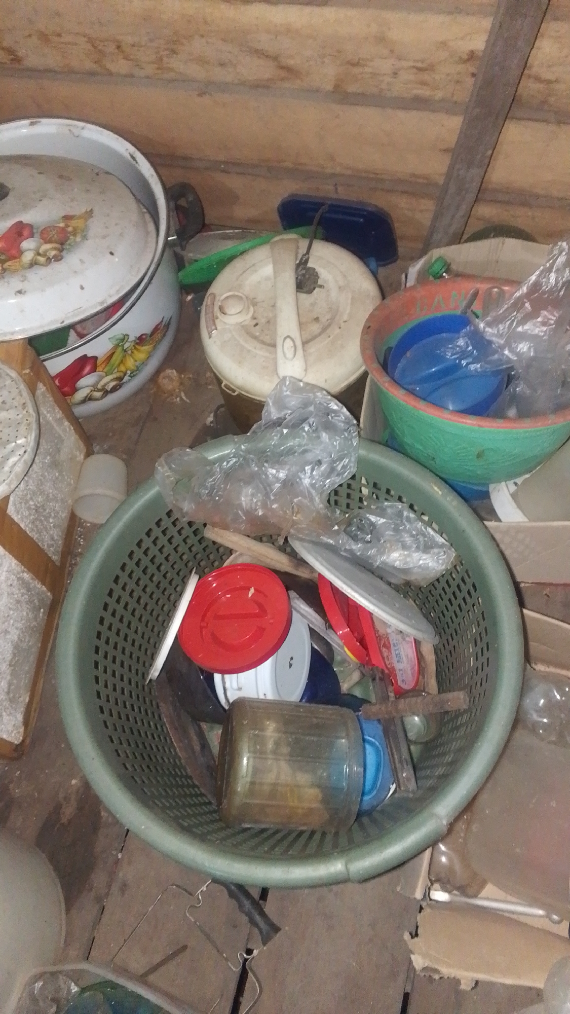 The image depicts a cluttered storage area, likely a shed or a room in a dwelling. The environment is rustic, with wooden plank walls and a wooden floor. Various household items are haphazardly stored.

In the foreground, a large green plastic basket is filled with objects. These include plastic containers, a large translucent plastic bottle with a yellow-brown liquid, red lids, white containers, and some plastic sheeting. A rusted metal object, possibly a tool or a piece of scrap metal, is also visible within the basket.

Behind the basket, several other items are visible. To the left, a large white pot with a decorative floral pattern on its lid is partially visible. Next to it, a white appliance with a cream-colored lid, possibly a rice cooker or a food processor, is present. A blue plastic container is placed behind this appliance. To the right of the appliance, a large, colorful bowl with green and orange rims contains blue plastic items and more plastic sheeting.

The overall impression is one of disorganization and storage of used or miscellaneous items in a non-ideal environment. There are no people visible in the image.

The provided location context, "Olak-Olak Kubu, Indonesia," suggests this scene is situated in a rural or semi-rural area of Indonesia. Given the rustic nature of the surroundings and the type of objects stored, it is plausible that this is a common storage method in such regions, prioritizing functionality and re-use of containers.