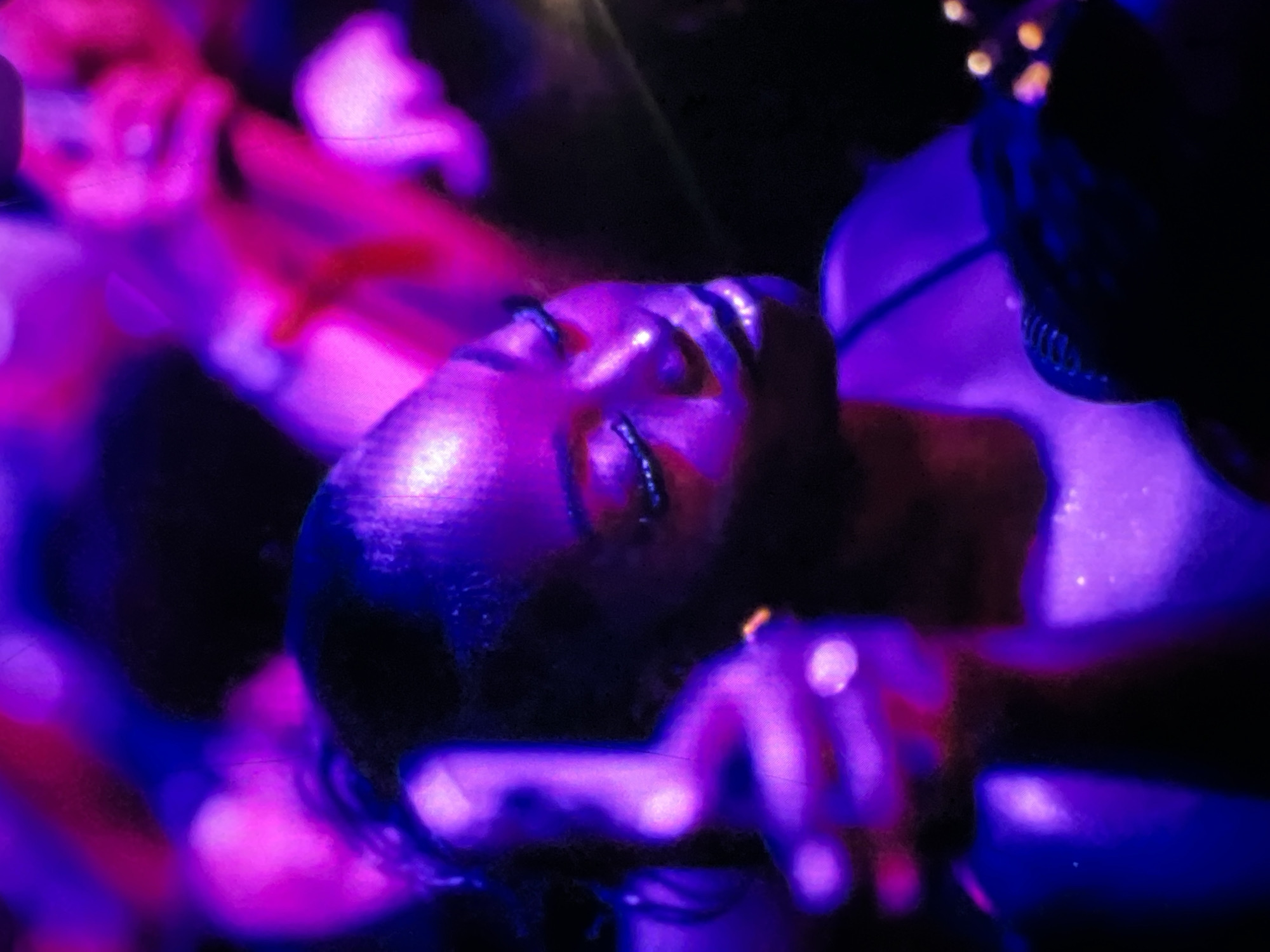 In Lagos, Nigeria, a woman is captured in a low-light, vibrant scene, illuminated by purple and pink lighting. She is in the foreground, her face turned to the side with her eyes closed and her lips parted slightly. Her makeup features glittery accents on her eyelids and a glossy finish on her lips. She appears to be holding a microphone or a similar object close to her face. In the background, other individuals are present, suggested by blurred shapes and vibrant lighting that indicates a lively environment, possibly a performance or a party. The overall atmosphere is energetic and immersive, typical of a night-time event in a bustling city like Lagos.