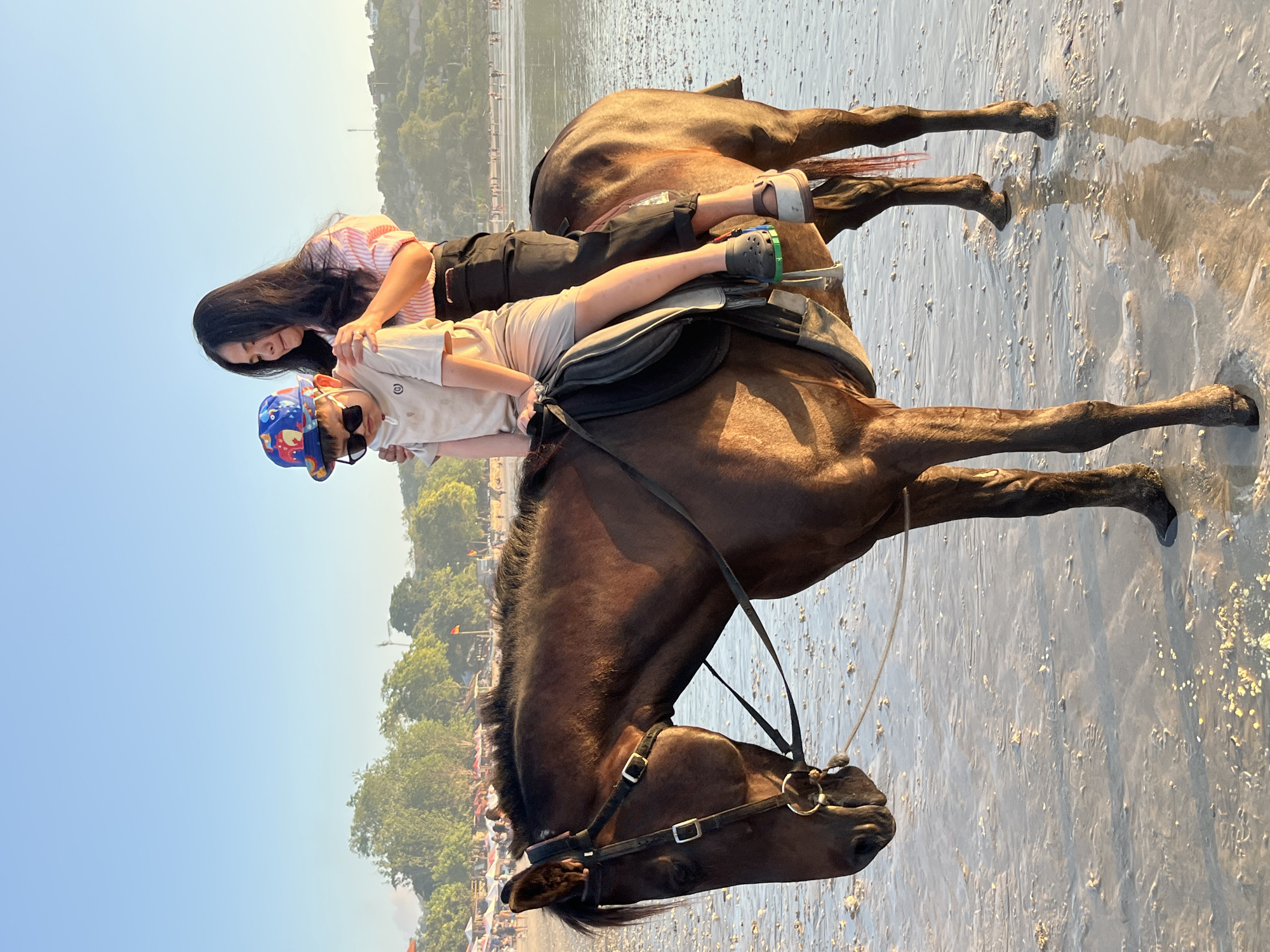 A woman and a child are riding a brown horse on the beach. The woman is behind the child, with her hands on the child's shoulders. The child is wearing a blue and red hat with an elephant print and sunglasses. The woman is wearing a pink and white striped shirt and black pants. The horse is wearing a bridle and saddle. The beach is wet sand, with some water on it. In the background, there are trees, people walking, and umbrellas. The sky is clear and blue. The location is the city of Kuta Selatan, Indonesia.