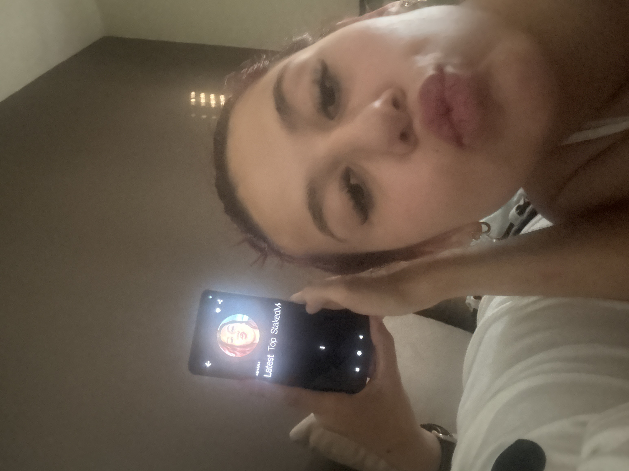A close-up selfie shows a young person, likely female, with dark hair, prominent eyebrows, and long eyelashes, making a playful "puckered lips" expression. Her face is tilted, and she is looking directly at the camera. She holds a black smartphone in her right hand, its screen brightly lit and facing the viewer.
The phone display shows what appears to be a social media or profile screen. There is a circular profile picture featuring a person with similar features to the individual taking the selfie, also making a puckered lips expression. Text on the screen includes "Latest Top StakedM" and a username that appears to be "ugcbaase". Other user interface elements like navigation arrows and dots are visible.
The person is wearing a light-colored top, possibly white, and a dark watch or bracelet on her left wrist. The setting is indoors, characterized by dim lighting. The background features a dark, solid-colored wall or surface, with several bright, small reflections or lights visible in the upper right, possibly from a distant chandelier or light fixture. The overall scene suggests a casual moment captured in low light.