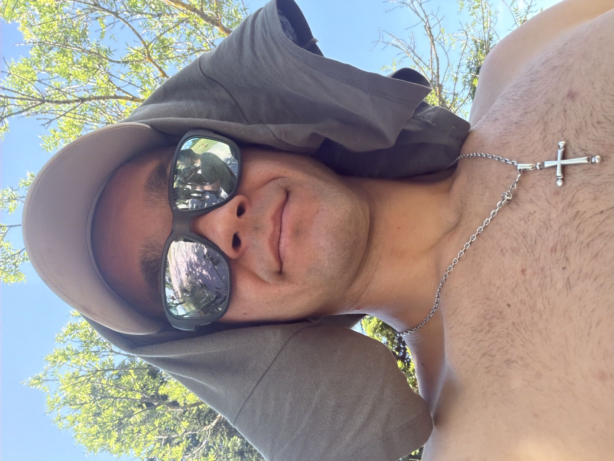 A man is taking a selfie outdoors on a sunny day. He is wearing a baseball cap and sunglasses, and a gray t-shirt is draped over his head and shoulders. A silver chain with a cross pendant is around his neck. The background shows green trees and a blue sky. The reflection in his sunglasses shows a path with people and trees. There is no indication of the location being Veliko Tarnovo, Bulgaria.