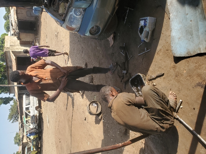 This image depicts an outdoor automotive repair scene in Kaduna South, Nigeria. The ground is unpaved earth. In the immediate foreground, a male individual in a dark brown, short-sleeved uniform squats barefoot, appearing to examine or work on a black metal pan filled with a dark liquid, possibly oil. Various hand tools, including wrenches and a long metal bar, are scattered around him. Centrally positioned, another male stands upright, wearing an orange short-sleeved button-up shirt and dark trousers, with his hands adjusting his lower front clothing. He wears sandals. To the right of the standing man, the front section of a light blue SUV is visible, its hood open, indicating ongoing maintenance. A white plastic fluid container and additional tools lie near the vehicle. In the mid-ground, several derelict white and yellow vans or minibuses are parked. In the background, a third male in a purple t-shirt walks away from the camera towards a cluster of single-story structures with corrugated metal roofs. The sky is bright and clear, and sparse vegetation is visible in the distance.