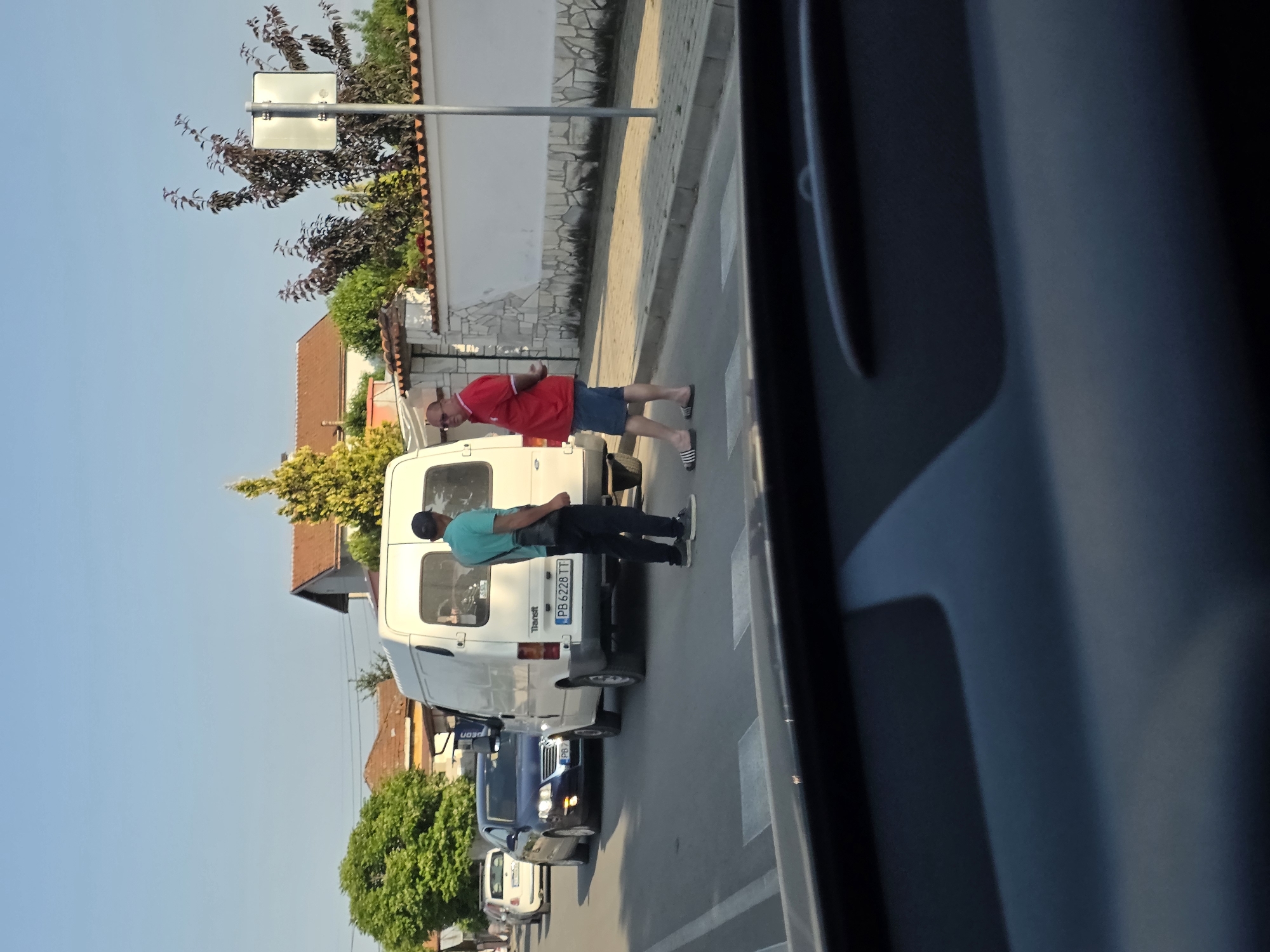 The image depicts a street scene in Yagodovo, Bulgaria, captured from within a vehicle. The foreground shows the dark dashboard of the observer's car. The midground features an asphalt road with visible pedestrian crossing markings. Two adult males are interacting on the right side of the road, adjacent to a white Ford Transit van (license plate PB 6228 TT) which is parked or stopped. The man on the left wears a light blue t-shirt, dark trousers, a dark cap, and a shoulder bag. The man on the right wears a red polo shirt, blue shorts, and black-and-white striped slides, with his hands clasped behind his back. Behind the van, a dark blue sedan (license plate PB 8...) and a white compact car are also present. A paved sidewalk runs along the right side of the road, bordered by a low white wall with decorative stone elements and terracotta tile coping. In the background, residential buildings with red-tiled roofs are visible amidst various trees and bushes, including a prominent bright green deciduous tree on the left and a yellow evergreen tree on the right. A light pole with a blank rectangular sign stands further down the street. The sky is clear and blue, indicating daytime conditions.