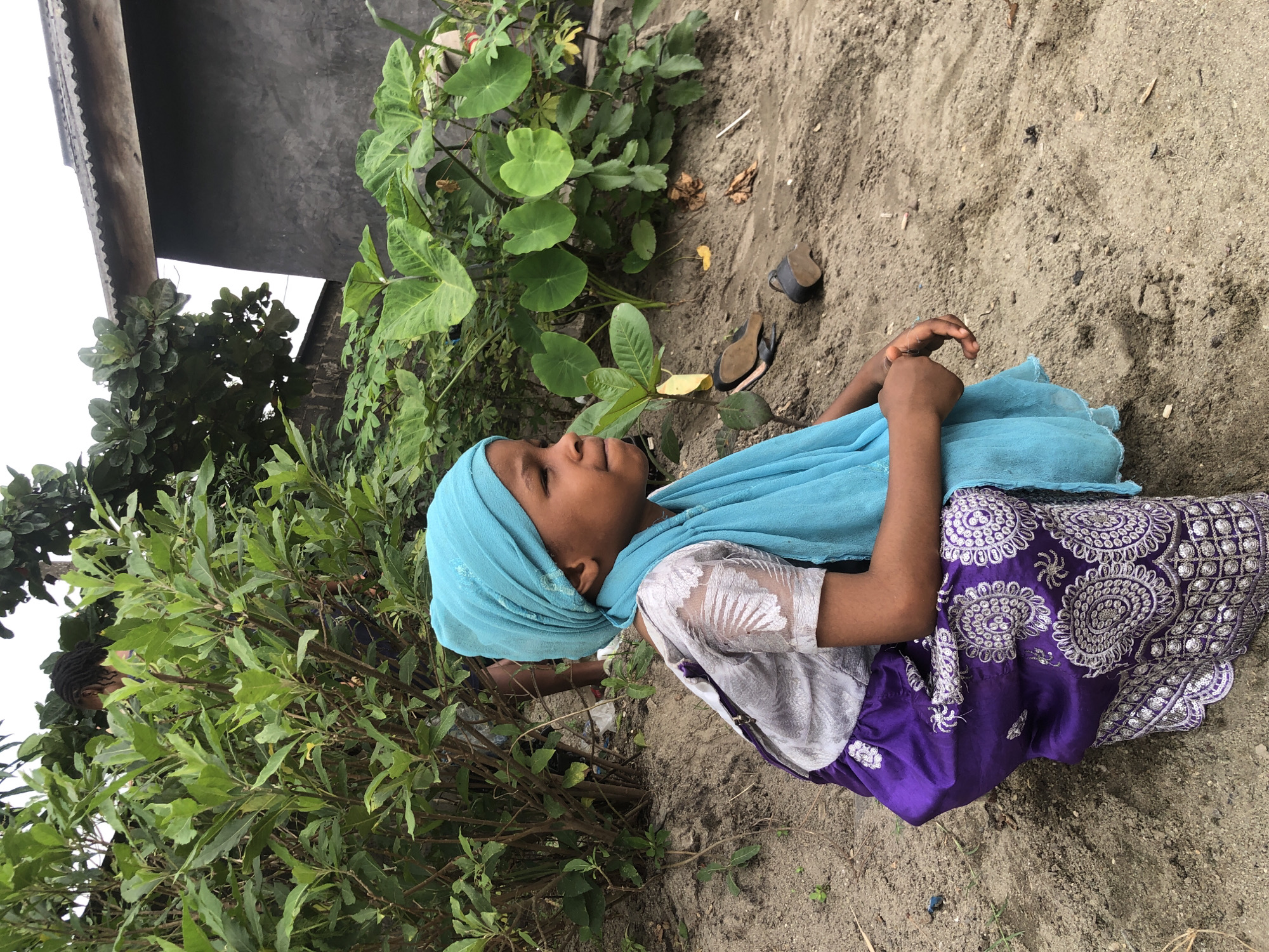 In Ado, Nigeria, a young girl is captured in a side profile, squatting on sandy ground amidst lush green foliage. She wears a vibrant turquoise headscarf and a matching scarf draped around her neck. Her attire includes a light-colored, possibly silver, blouse with intricate patterns on the sleeves and a deep purple skirt adorned with white circular embroidered designs and sequins. Her hands are clasped in her lap, and she gazes thoughtfully to her right.
Behind her, a dense green bush forms a natural backdrop, with its branches reaching upwards. To the right of the frame, a portion of a building with a corrugated iron roof and a dark grey concrete wall is visible, suggesting an urban or semi-urban setting. Scattered on the sandy ground are a pair of dark sandals and what appears to be a small black object, possibly a container or shoe heel. The ground itself is a mix of sand and dry leaves, indicating an outdoor environment. A person's legs and feet, partially obscured by the foliage, are visible in the background. The lighting suggests an overcast day.