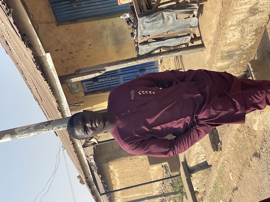 A young adult male of dark complexion, wearing a maroon traditional two-piece outfit with white chevron embroidery on the left chest pocket, stands centrally in an outdoor setting in Unguwar Gabas, Nigeria. He faces the camera directly, hands in his pockets. A utility pole is situated behind his head. The background features a faded yellow single-story building with a corrugated metal roof and blue metal doors/shutters. To the subject's right, a wooden table displays various items including sandals and a water bottle, with corrugated metal sheets leaning beneath it. The ground consists of dirt with scattered debris, and concrete steps or a raised curb are visible in the foreground. The scene is brightly illuminated by daylight.