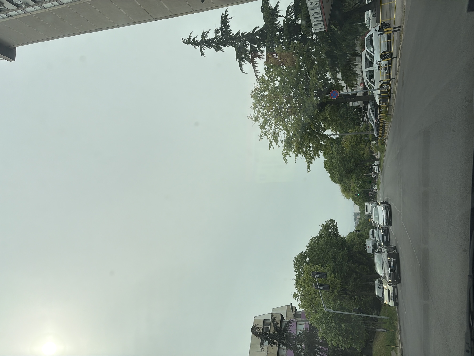 The image depicts an urban or suburban street scene under an overcast sky, with a bright spot in the lower left corner possibly indicating the sun or a strong reflection. The perspective appears to be from inside a vehicle, looking towards a road lined with trees and buildings.
On the far left, a large, modern building structure with a concrete overhang is prominently visible. The right side of the image features a multi-lane asphalt road with cars. Several white vehicles are parked along the right curb, separated from the main road by yellow barriers. Behind these parked cars, a building sign partially reads "HOTELS," framed by lush green trees, including tall conifer-like varieties and broader deciduous trees. A blue circular road sign, likely indicating "no entry" or "forbidden," is also visible near the "HOTELS" sign.
Further down the road, a line of cars is stopped in traffic. Notable vehicles in the queue include a black Alfa Romeo with the license plate "BG 2712-KX" and a white Smart car. Another vehicle immediately behind the Alfa Romeo has the license plate "BG 0244-GX." A green traffic light is visible in the distance, indicating that the traffic is likely waiting for the signal to change. The road is bordered by dense foliage, and another building with distinct purple, pink, and white sections can be seen in the distance on the left side of the road.
The "BG" prefix on the license plates confirms the location as Belgrade, Serbia, consistent with the provided context of Vozdovac Urban Municipality. The scene captures a typical daytime urban environment with traffic and mixed-use buildings.