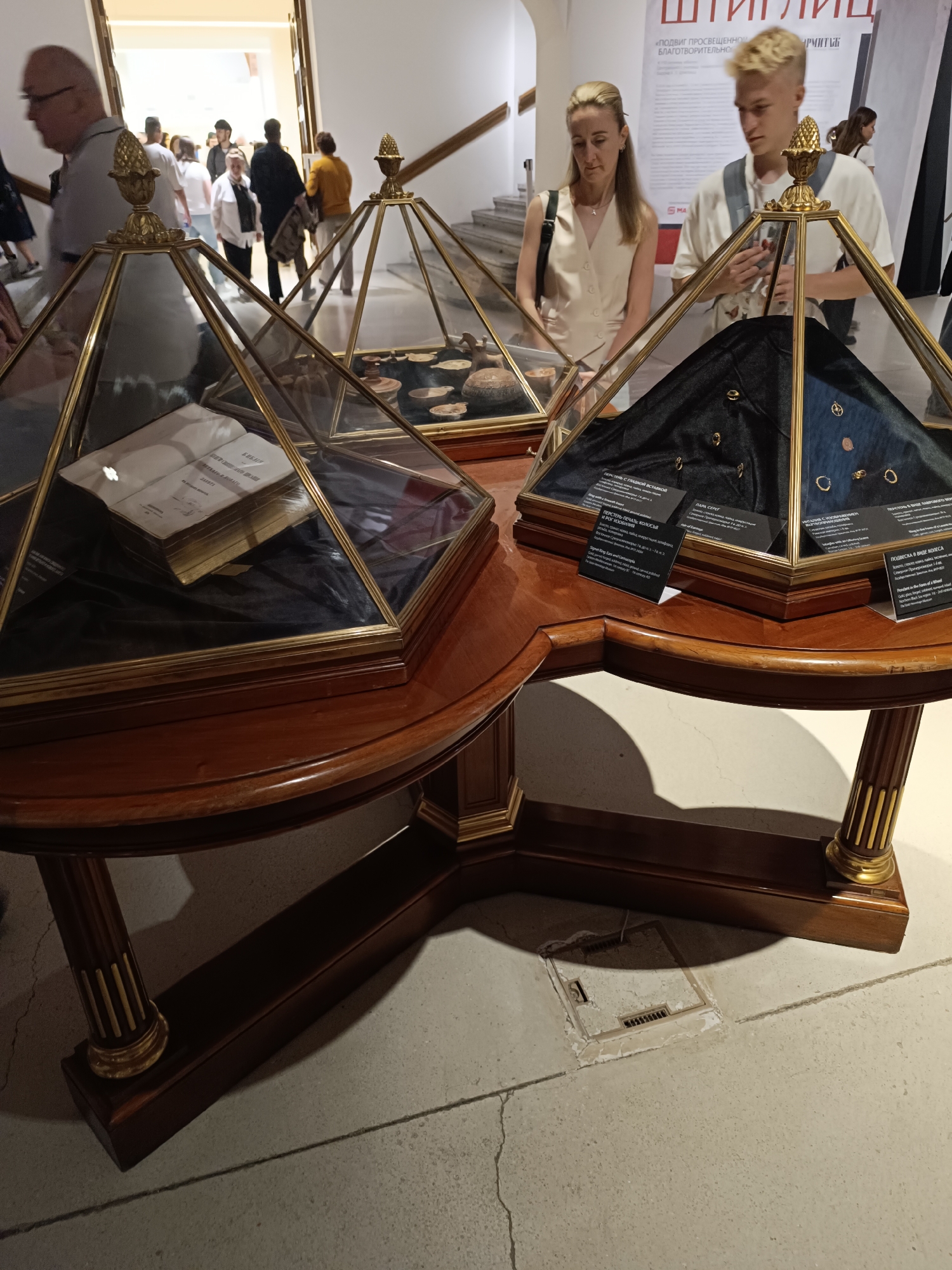 A museum exhibit is shown, featuring historical artifacts displayed under glass domes on a wooden table. A book is visible in one dome, and jewelry and pottery are in another. Several people are present, including a woman and a young man examining the exhibit, and other visitors in the background. The setting appears to be an indoor public space with stairs and signage.