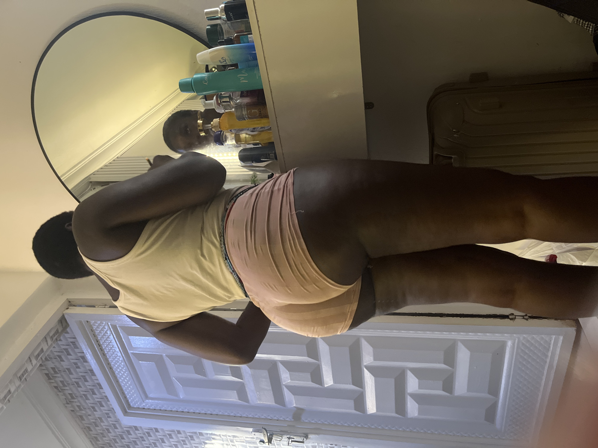 A person is standing in a room, facing a mirror and a dresser. The person is wearing a tank top and shorts. The dresser has various bottles and containers on it. There is a door to the left of the frame. The room appears to be a bedroom or dressing area. The image does not provide enough information to determine the specific location within Abuja, Nigeria.