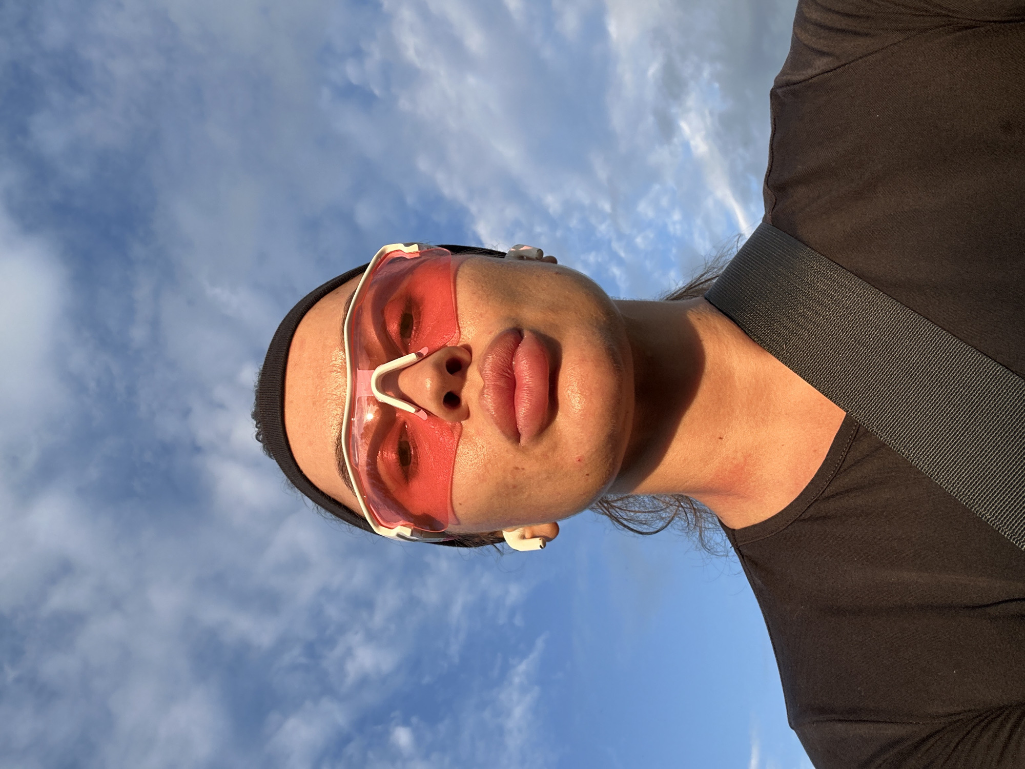 A woman is pictured from the chest up, outdoors, with a sky full of clouds in the background. She is wearing red-tinted sunglasses, a black headband, and a black t-shirt. There is a gray strap across her chest. She has white earbuds in her ears. The woman's skin appears slightly red on her neck. The overall lighting suggests it is either early morning or late afternoon. No specific Yuzhnouralsk, Russia landmarks are visible.
