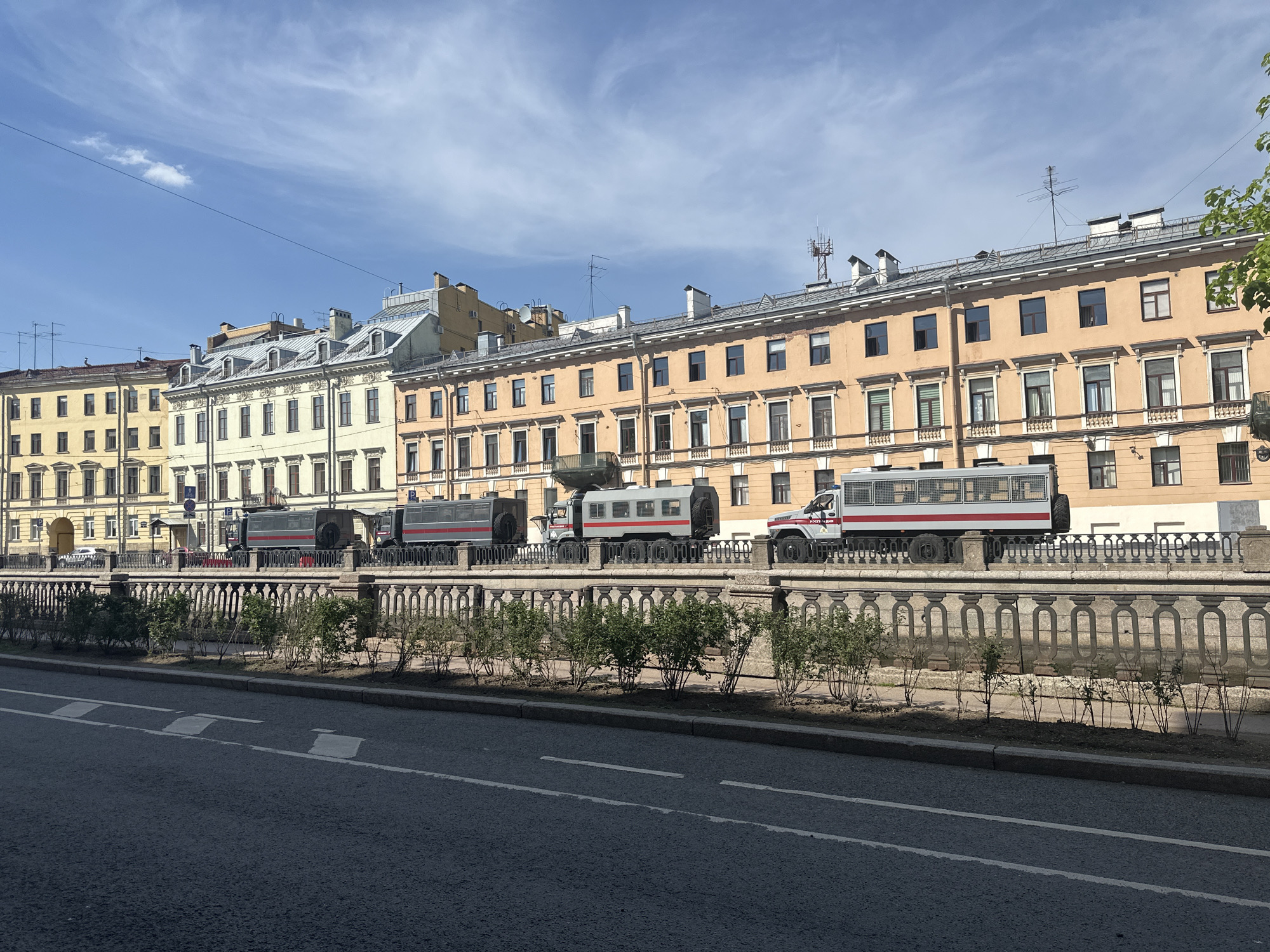 The image depicts a street scene in Saint Petersburg, Russia, with several large military-style vehicles parked along a canal or river embankment. The vehicles, which are predominantly dark gray and silver with red stripes, are parked parallel to the road and are near a stone balustrade. Behind the balustrade, there are rows of buildings with classical architectural features. The buildings are a mix of yellow and beige colors and have multiple windows and decorative elements. A row of small bushes lines the embankment. The foreground shows the paved surface of a street with painted lane markings. The sky is blue with scattered clouds. There are no people visible in the frame.