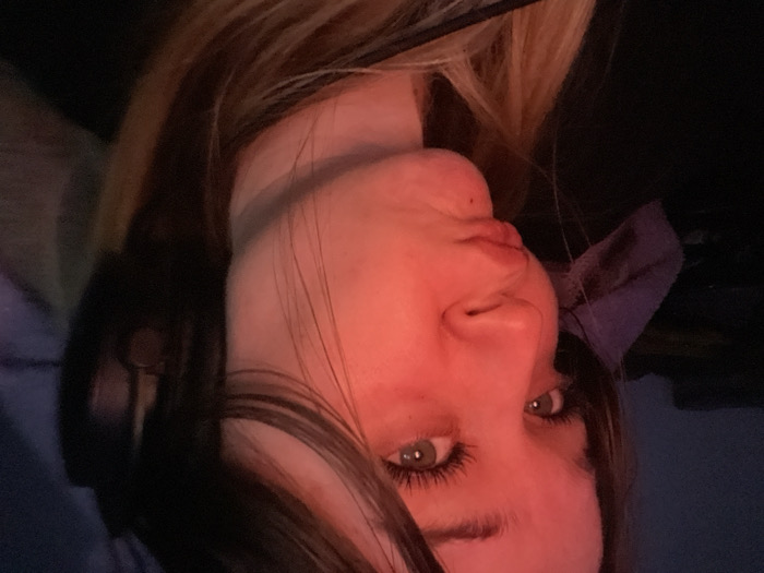 A young female subject is depicted indoors in the city of Pushchino, Russia. Her face, captured in a close-up, is strongly illuminated by a red light source from the left side of the frame, casting a reddish hue across her skin. She has long, dark hair with lighter strands visible at the bottom right. Her visible left eye is green and features prominent eyelashes. Her lips are pursed in a pout, and she maintains a direct gaze towards the camera. Dark over-ear headphones are worn on the right side of her head, partially visible. A small dark mark is present on her chin. The background is dark, out of focus, and indistinct, with a light purple fabric item visible on the far left.