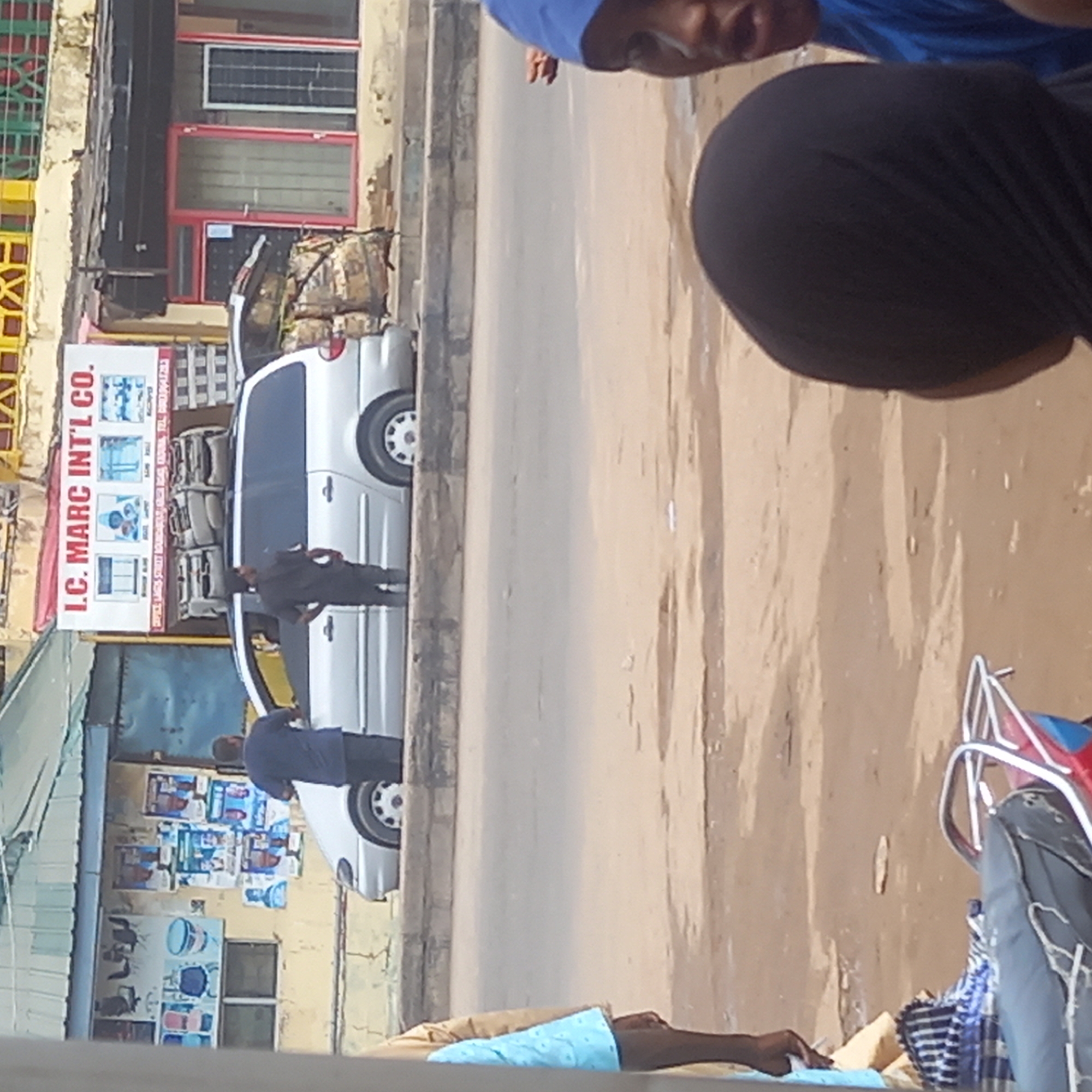 The image, taken in Sardauna, Nigeria, depicts a street scene. A white minivan is parked along the side of the road. Two men are standing near the van. One is wearing a dark jacket and the other a blue shirt. A sign for "I.C. MARC INT'L CO." is visible on a building. Posters are plastered on a wall near the minivan. In the foreground, parts of people, possibly vendors, are visible with merchandise. The road is dusty. Buildings with shops are visible in the background.