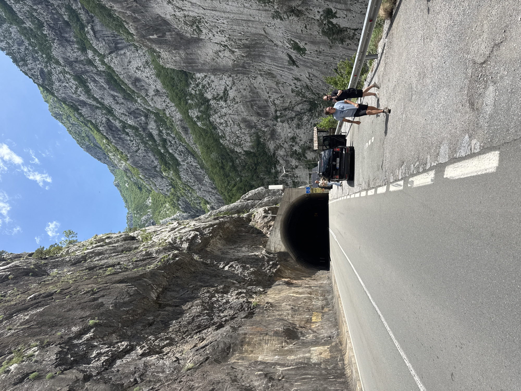 The image depicts a road winding through a mountainous region, possibly near Vlahovići, Montenegro. A dark tunnel entrance is visible in the center-left of the image. A black car is parked near the tunnel entrance, with its door open. Several individuals stand near the car, possibly interacting. Two people are standing on the right side of the road, seemingly observing the scene. The environment consists of towering rocky mountains, one with sparse vegetation and the other is rocky with green vegetation on the upper left side of the frame. The sky is visible in the top left corner and is light blue with a few clouds.