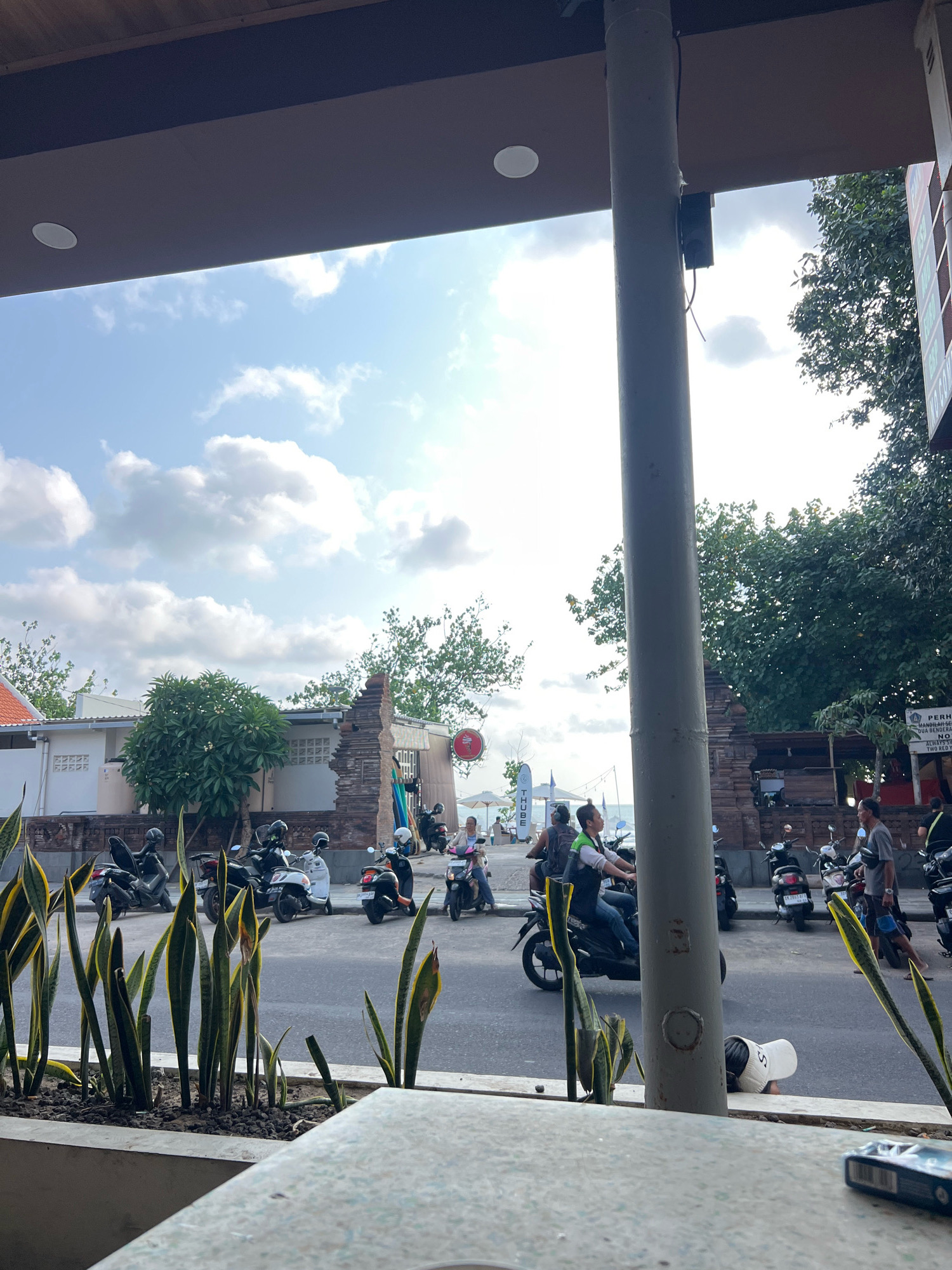 The image depicts a street scene in Kuta, Indonesia, likely near a beach access point, with a view of the sky and some buildings. In the foreground, a concrete planter contains several snake plants. A large, grey cylindrical pole is prominently positioned in the middle ground, obscuring parts of the scene.
The street is lined with parked scooters and motorcycles, with several people riding them. Some individuals are wearing helmets. On the right side of the street, a sign with text is visible. In the background, there are trees, a building with an orange roof, and what appears to be a beach with umbrellas and people in the distance. The sky is blue with scattered clouds. The overall atmosphere suggests a bustling, tropical environment.