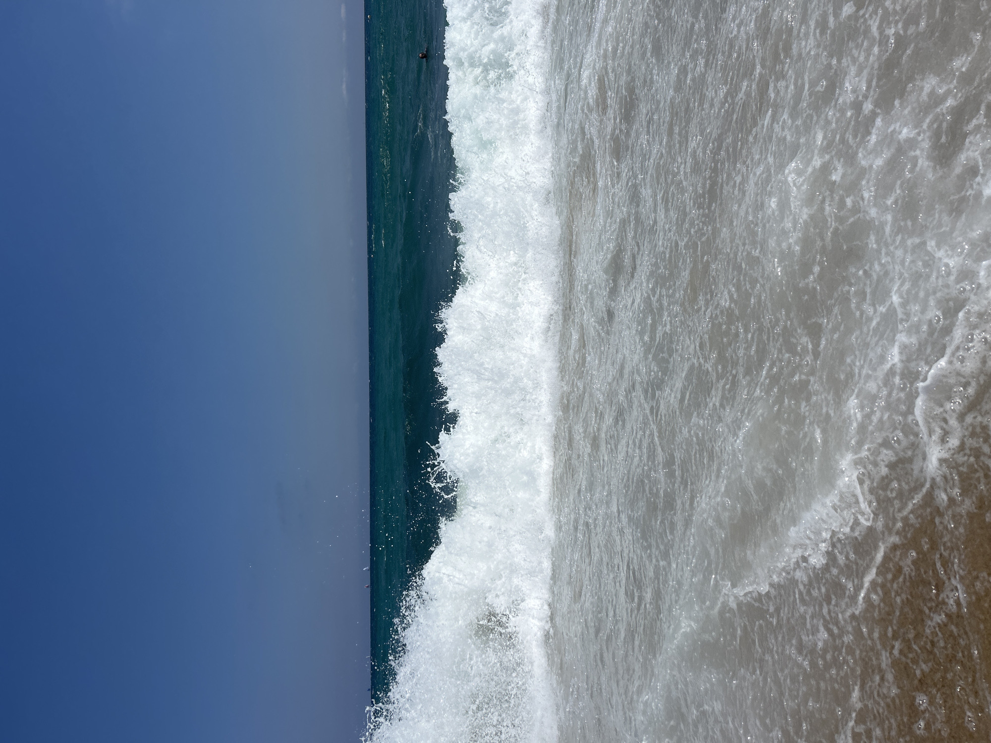 A wide shot of a beach in Barcelona, Spain shows a large wave crashing on the shore. The water is a vibrant teal and the sky is a clear, deep blue. There are a few sailboats in the distance and a person swimming in the ocean. The sand is light brown.
