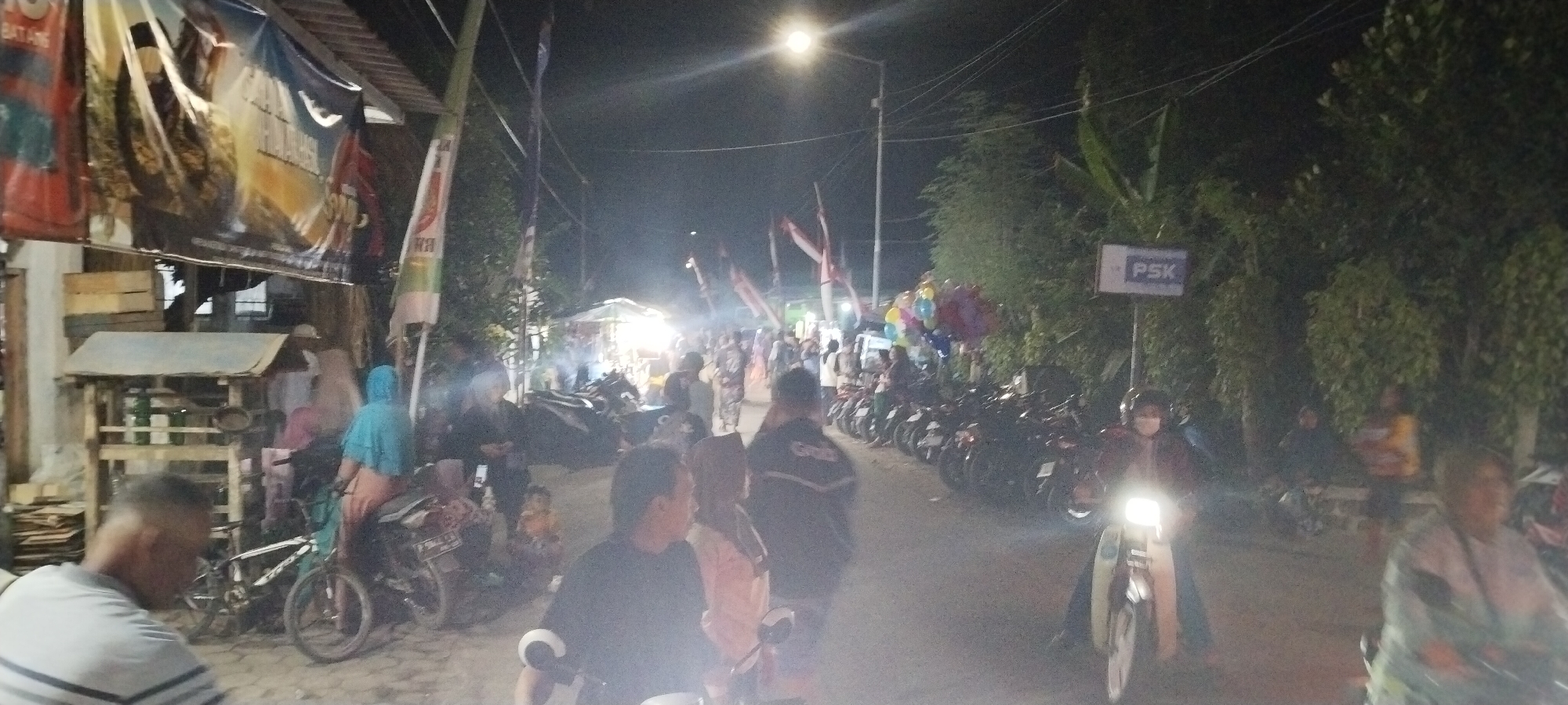 The image depicts a nighttime street scene in Keting, Indonesia, likely during a local gathering or event. The street is lined with trees and has a row of parked motorcycles on the right side. A sign with the letters "PSK" is visible on the right. Several people are present, some on motorcycles, others standing or walking. Banners are hung across the street, and there are lights illuminating the area, suggesting a festive atmosphere. In the foreground, a man wearing a striped shirt is partially visible, facing away from the camera. Further back, individuals are seen on motorcycles, including one with its headlights on, and others are gathered in groups. The overall impression is a busy, outdoor community event at night.