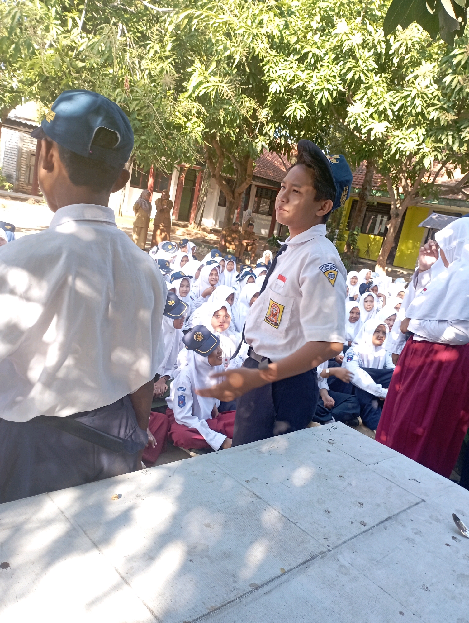 In Banyuurip, Indonesia, a group of students, primarily girls in white uniforms and hijabs, are seated outdoors under the shade of leafy trees. Some students are looking towards the foreground, while others are engaged with each other. In the foreground, two male students in blue caps and white shirts are visible. One is facing away from the camera, his back to the viewer, while the other is turned to the side, looking upwards and to his left. A light-colored, rectangular surface, possibly a table, is in the immediate foreground, with some debris and shadows on it. The environment suggests a school setting, likely during an outdoor activity or assembly.