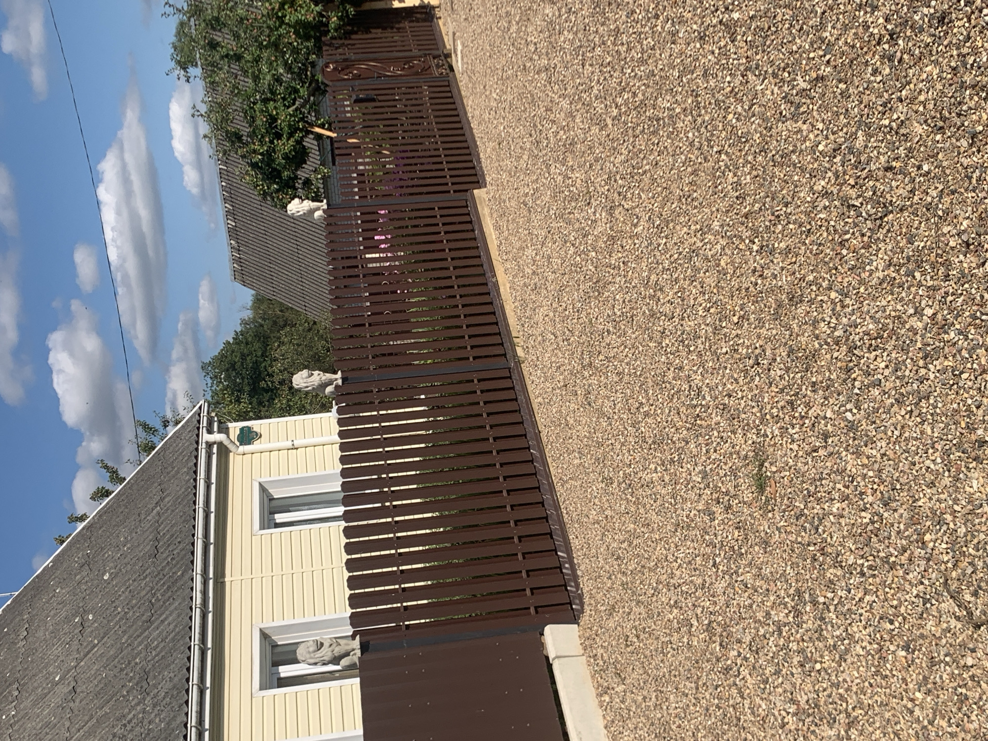 The image shows a gravel driveway in front of a residential property. A brown metal fence with vertical slats runs across the front of the property. Two stone lion statues are visible on either side of the fence posts near the house. The house has light yellow siding and a grey, corrugated roof. The sky is blue with scattered white clouds. The location is in the city of Kapyĺ, Belarus.